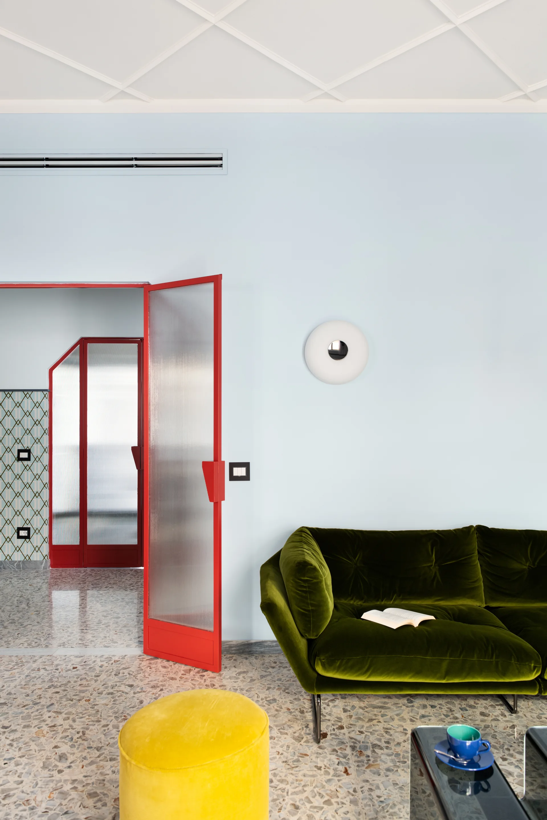 A modern living room features a green velvet sofa, a yellow velvet ottoman, and a red-framed glass door against light blue walls.
