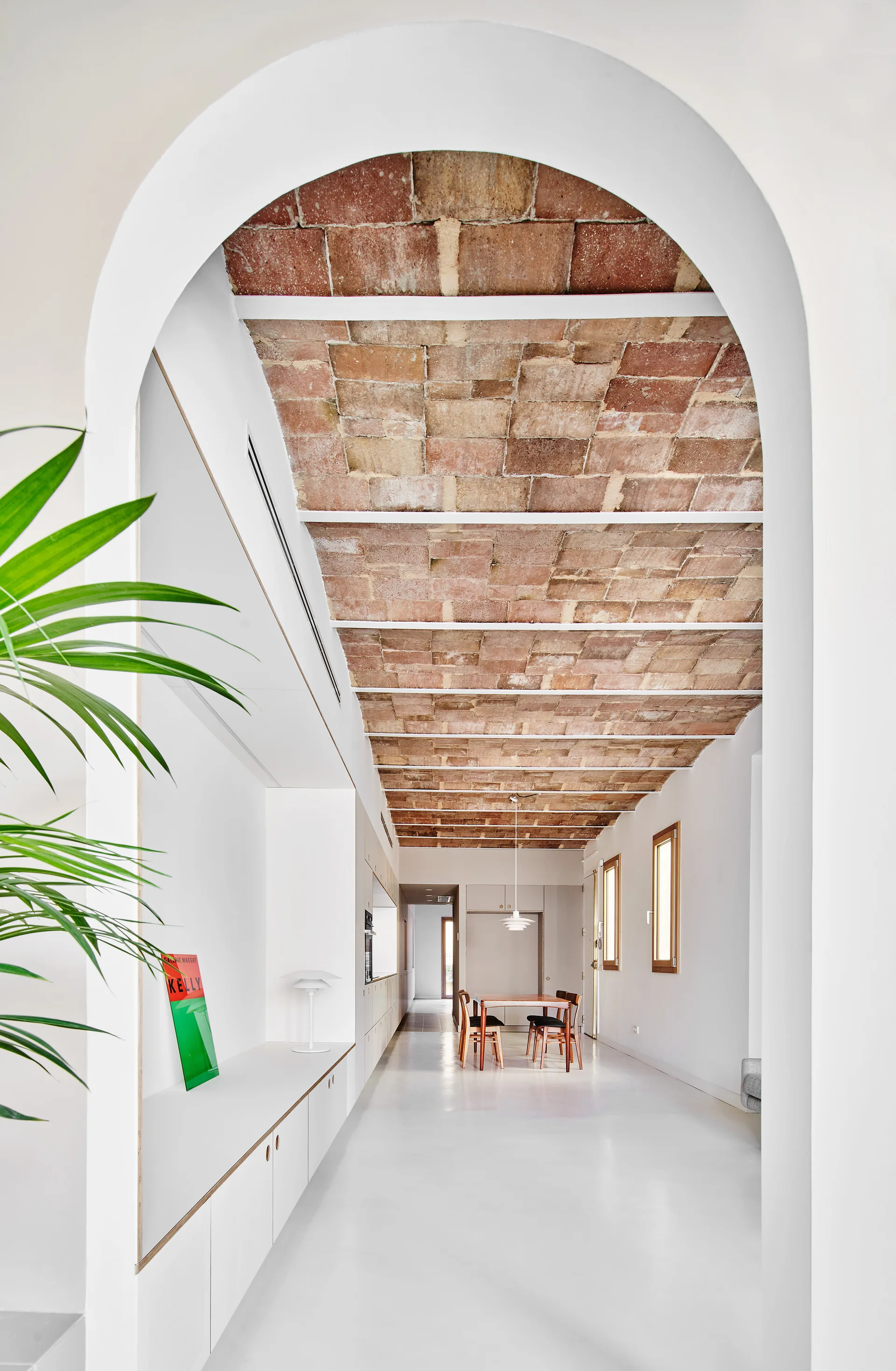 A view through a white plastered archway into a long minimalist room featuring a vaulted brick ceiling, smooth white flooring, and a distant wooden dining set.