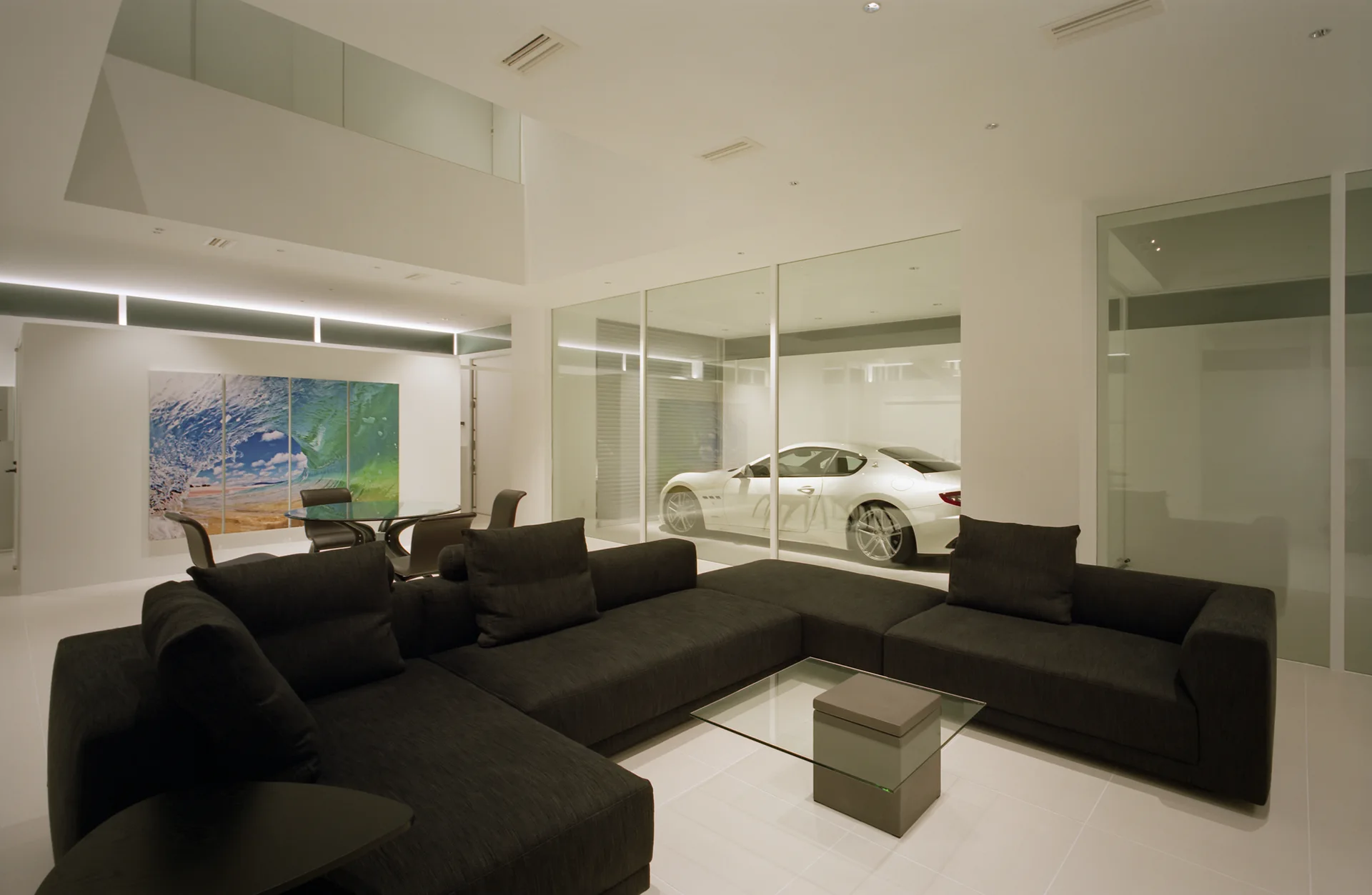 A large charcoal sectional sofa sits on a white tiled floor in a minimalist living room featuring a glass wall looking into a garage with a white car.