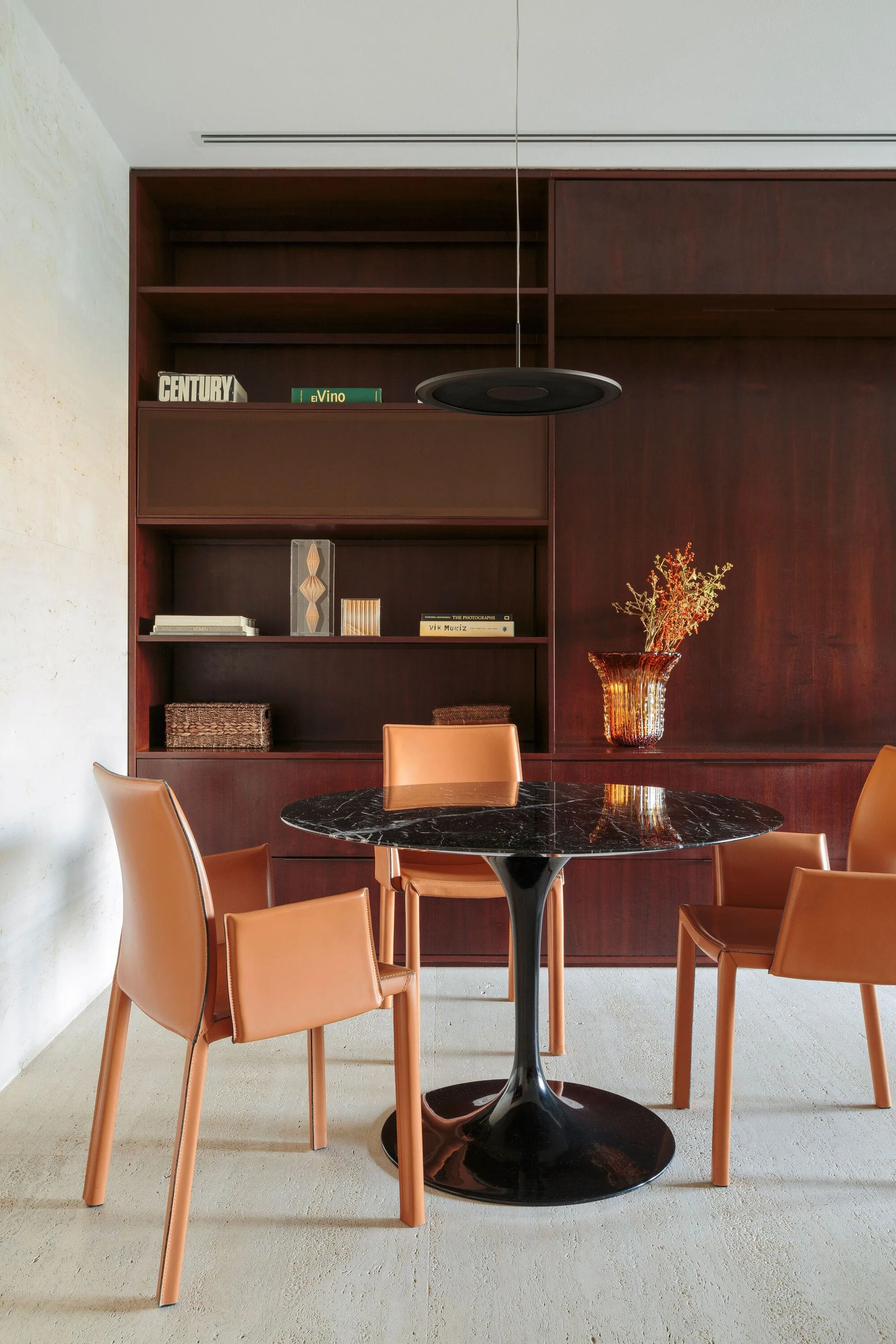 A round black marble pedestal dining table is surrounded by three cognac leather armchairs in front of a dark brown wood built-in shelving unit.