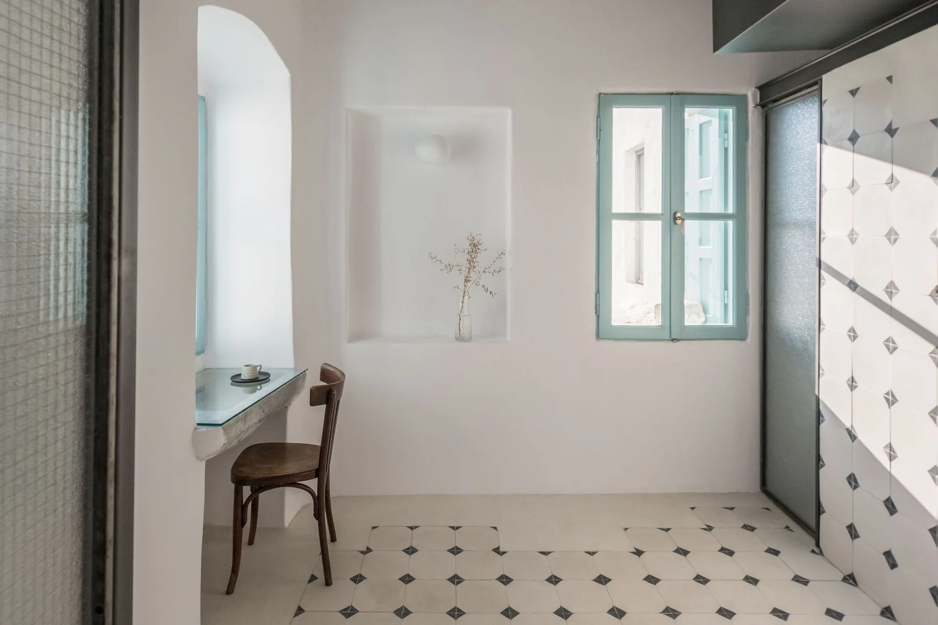 A bright white room features a built-in plaster desk with a wooden chair, a blue-framed window, and geometric diamond-patterned floor tiles.