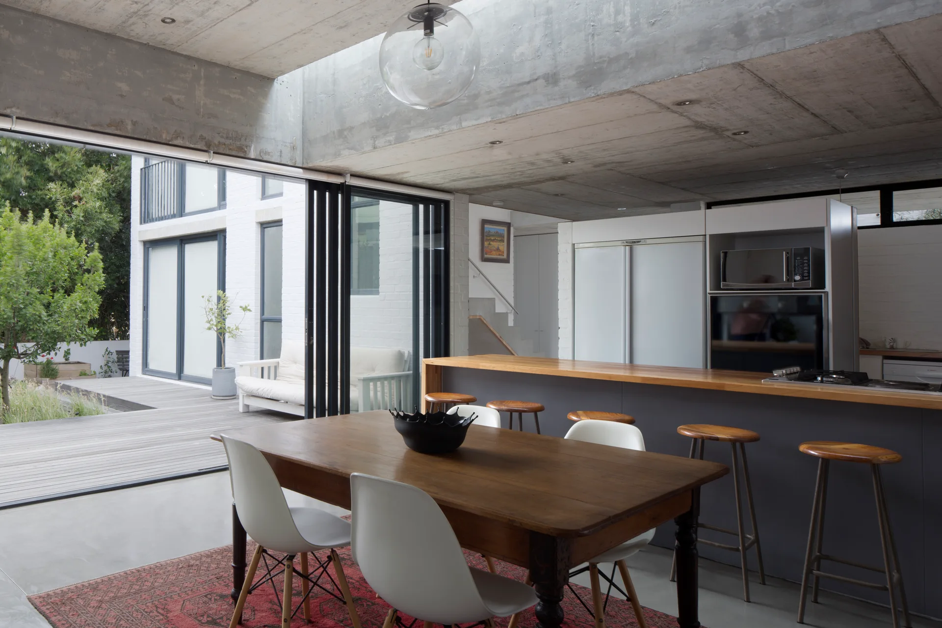 An open dining room features a wooden table, molded white chairs, and an oriental rug under an exposed concrete ceiling.
