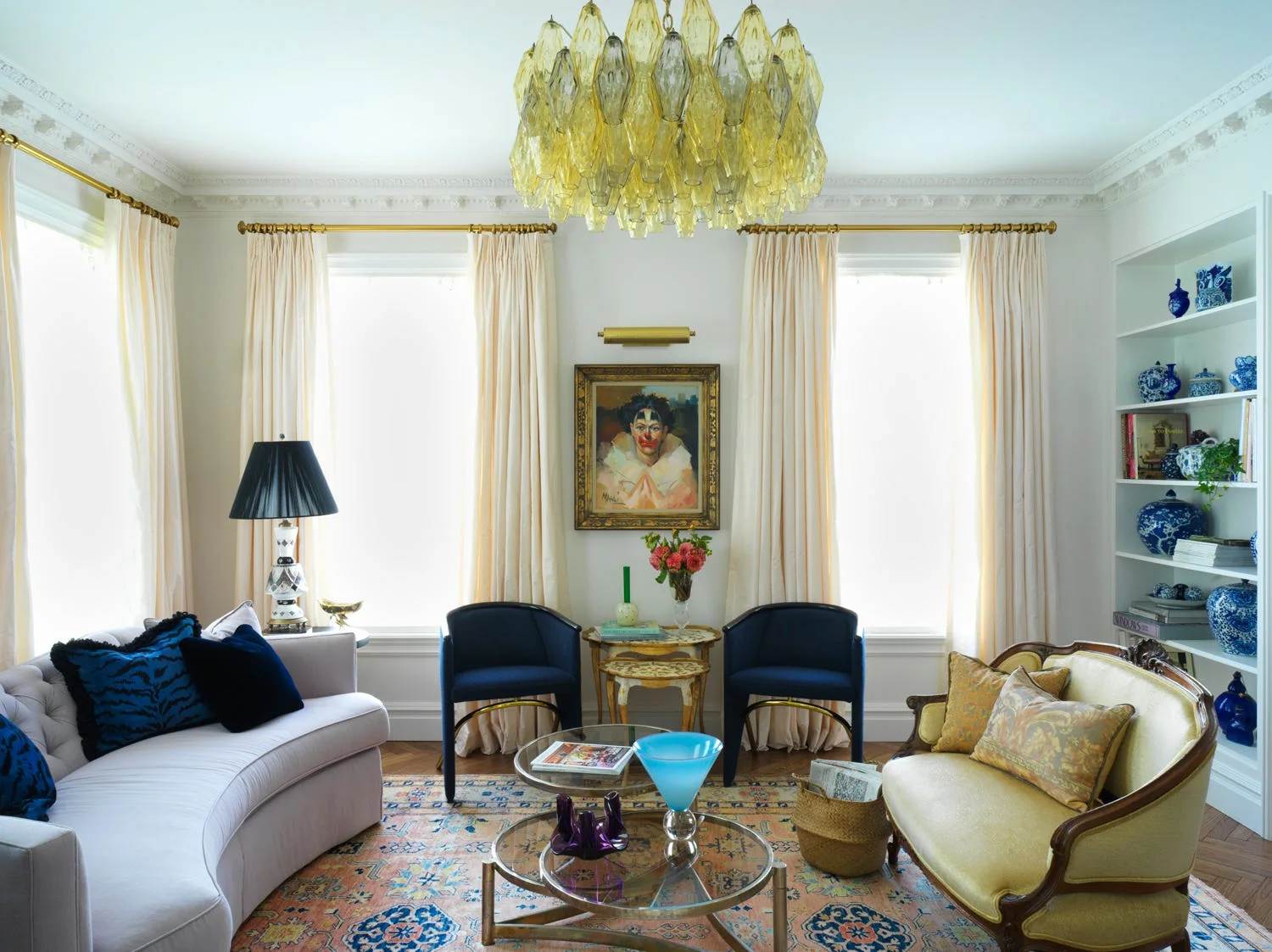 A living room features a yellow tiered glass chandelier, curved light pink sofa, two navy armchairs, and white built-in bookshelves.