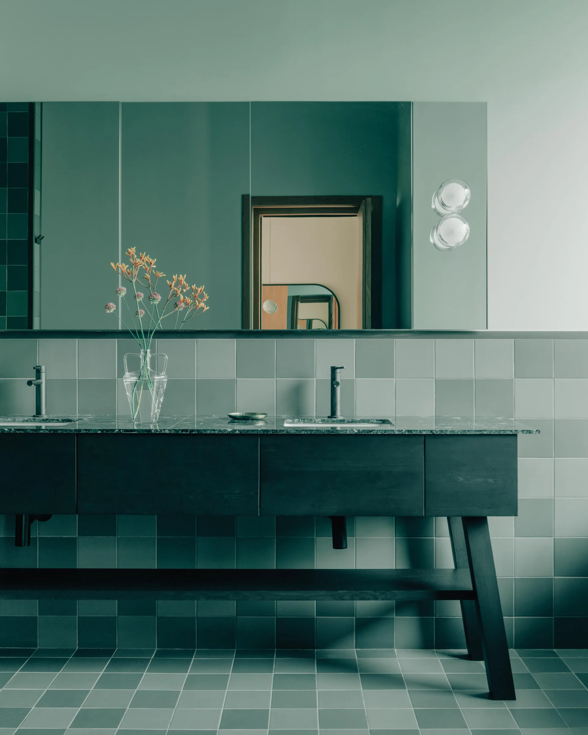 A modern bathroom features a dark wood double vanity under a large mirror, surrounded by sage green square wall and floor tiles.