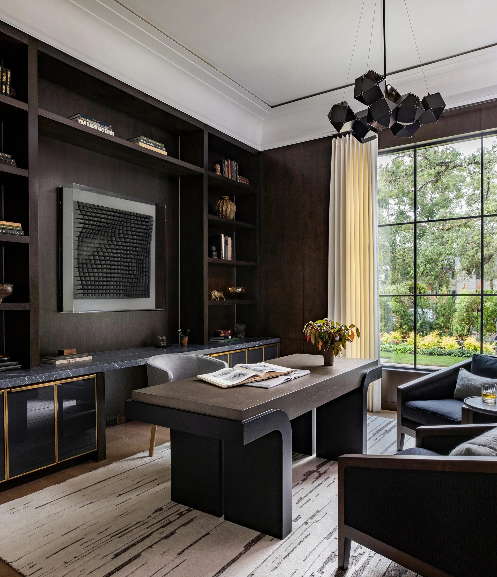 A home office with dark wood built-in bookcases, a thick modern desk, a geometric black chandelier, and a grid window looking out at trees.