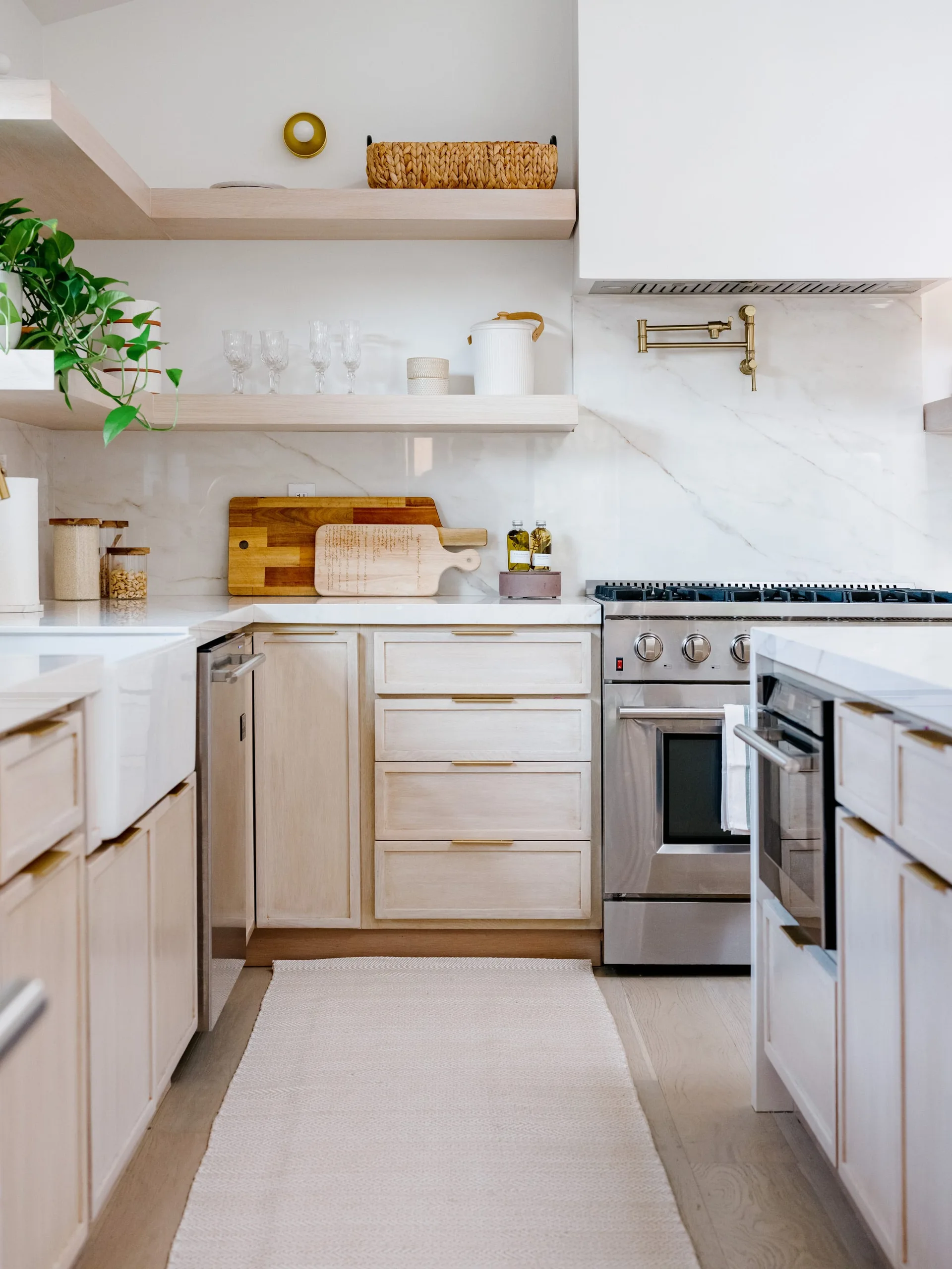 A kitchen featuring light wood lower cabinets, a white marble backsplash, floating shelves, and a stainless steel stove with a brass pot filler.
