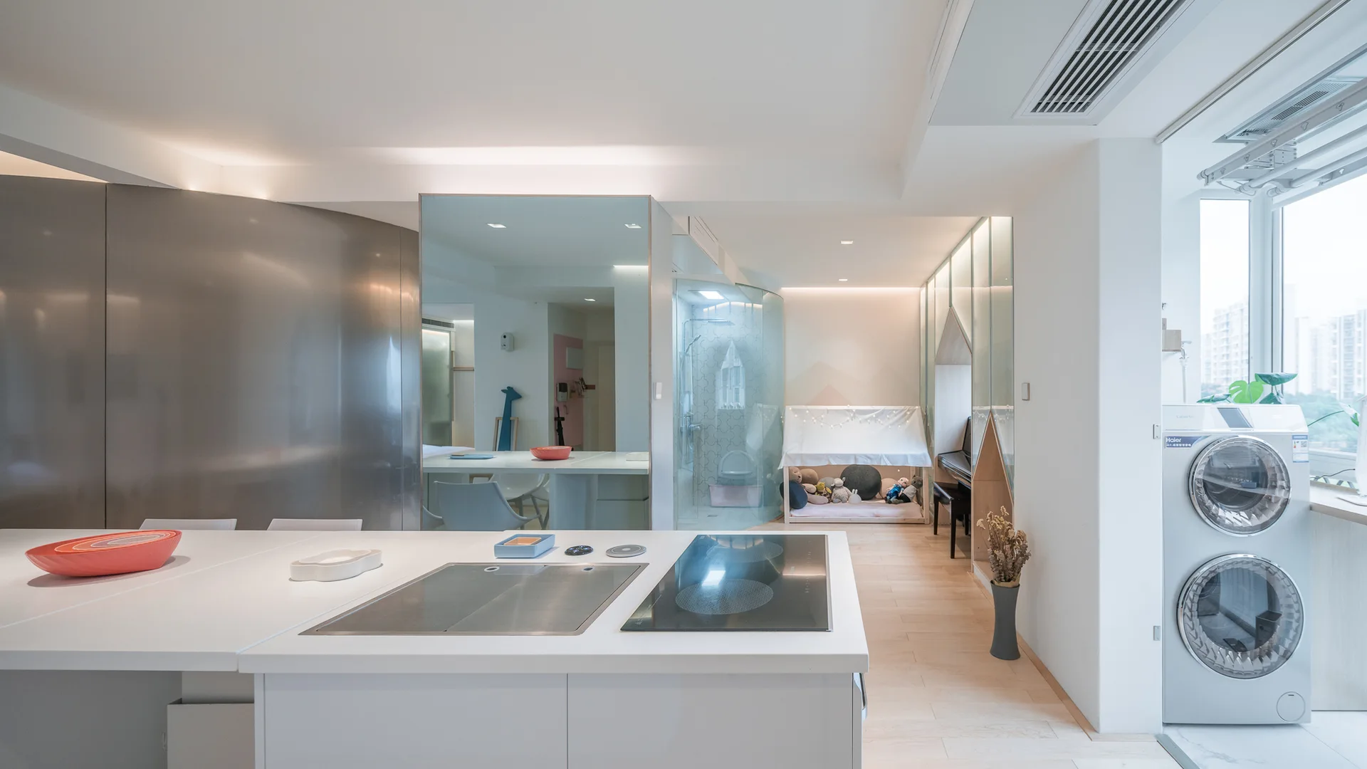 A modern white kitchen island faces a multi-functional room with a curved glass bathroom wall, a large mirror, a kids' play tent, and stacked laundry.