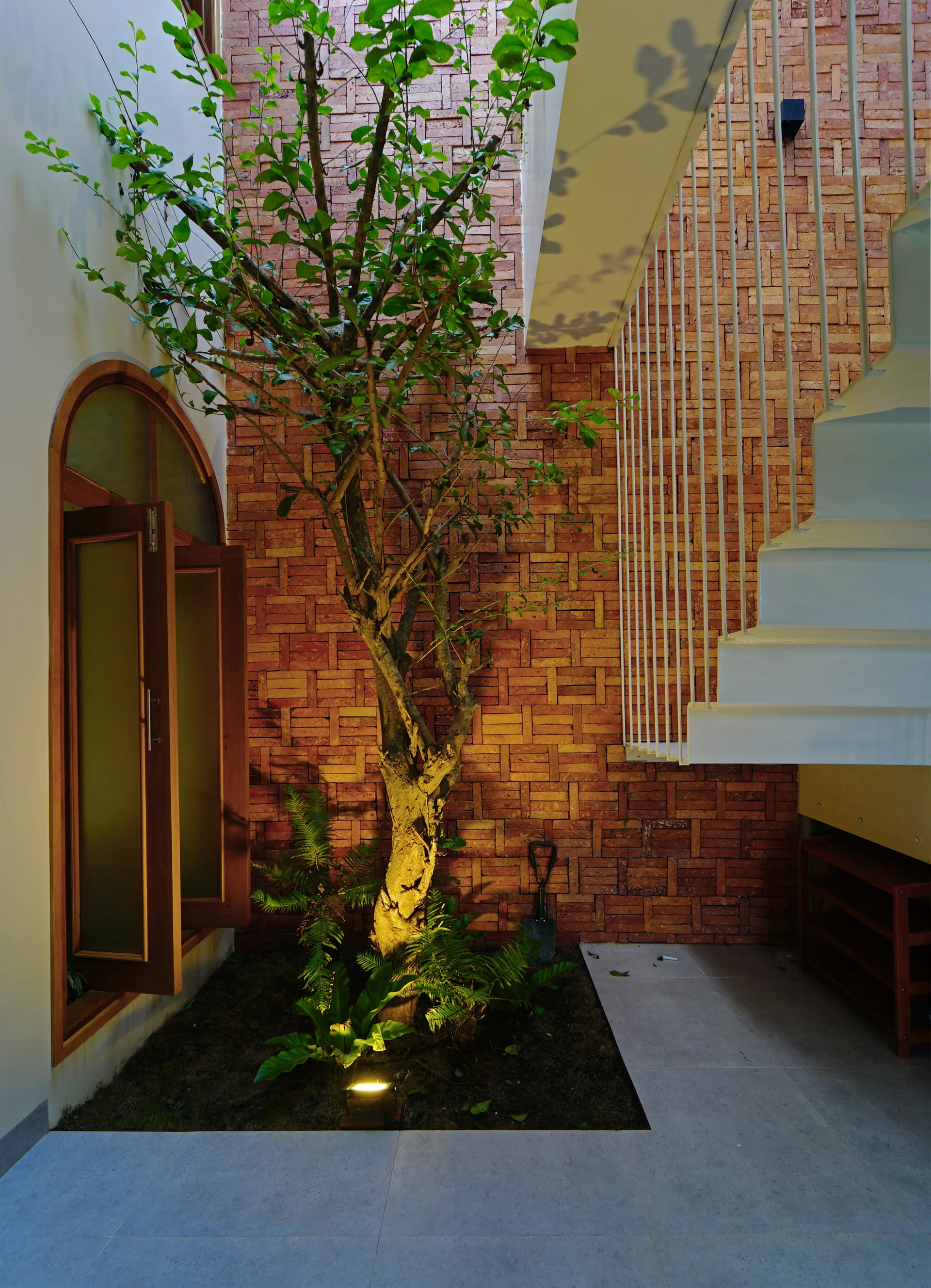 An indoor courtyard features a slender green tree planted in a soil bed, positioned against a textured red brick wall next to a white metal staircase.