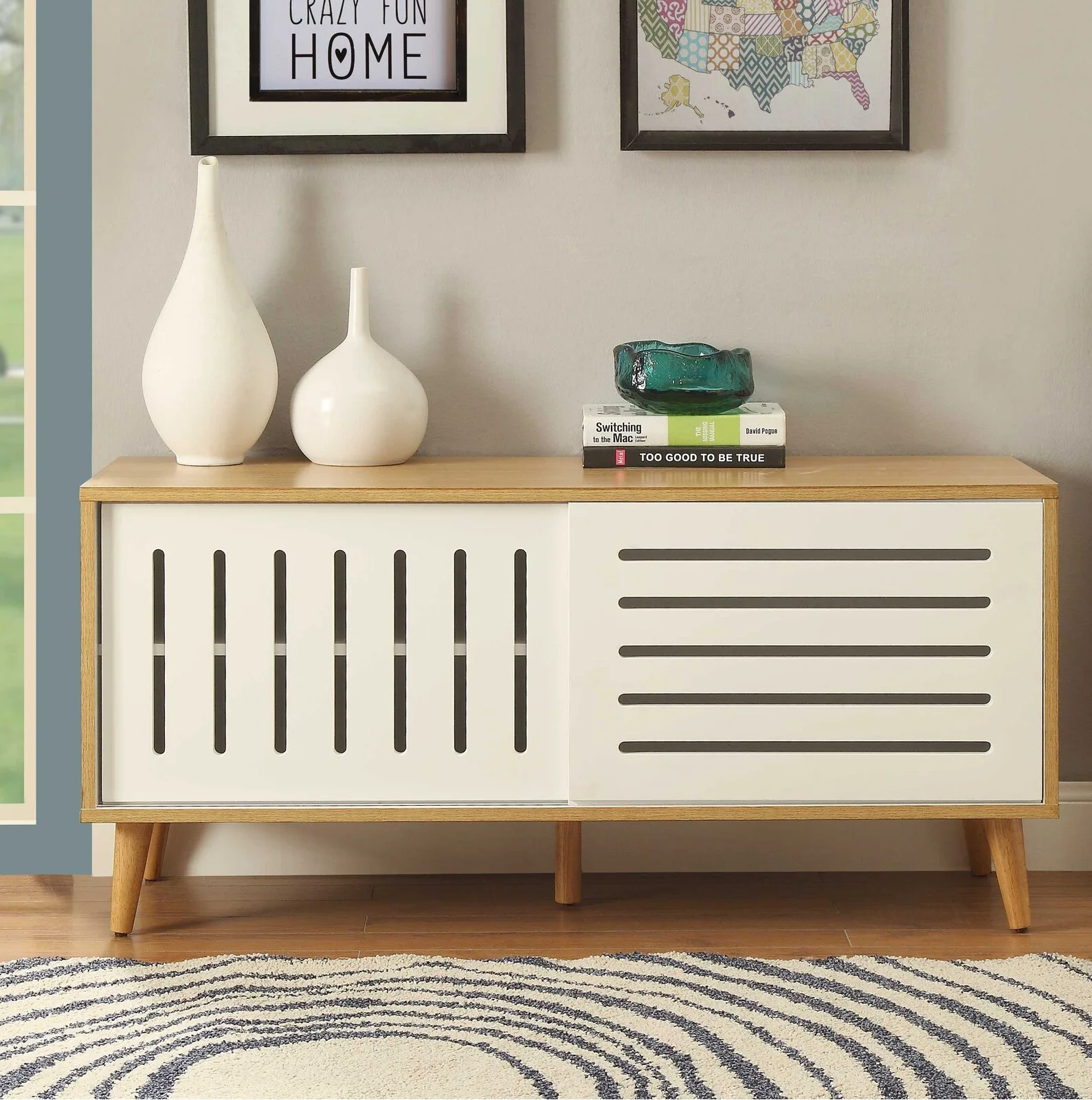 A low wooden cabinet with white sliding slatted doors sits on a wood floor with a patterned rug beneath ceramic vases and framed art.