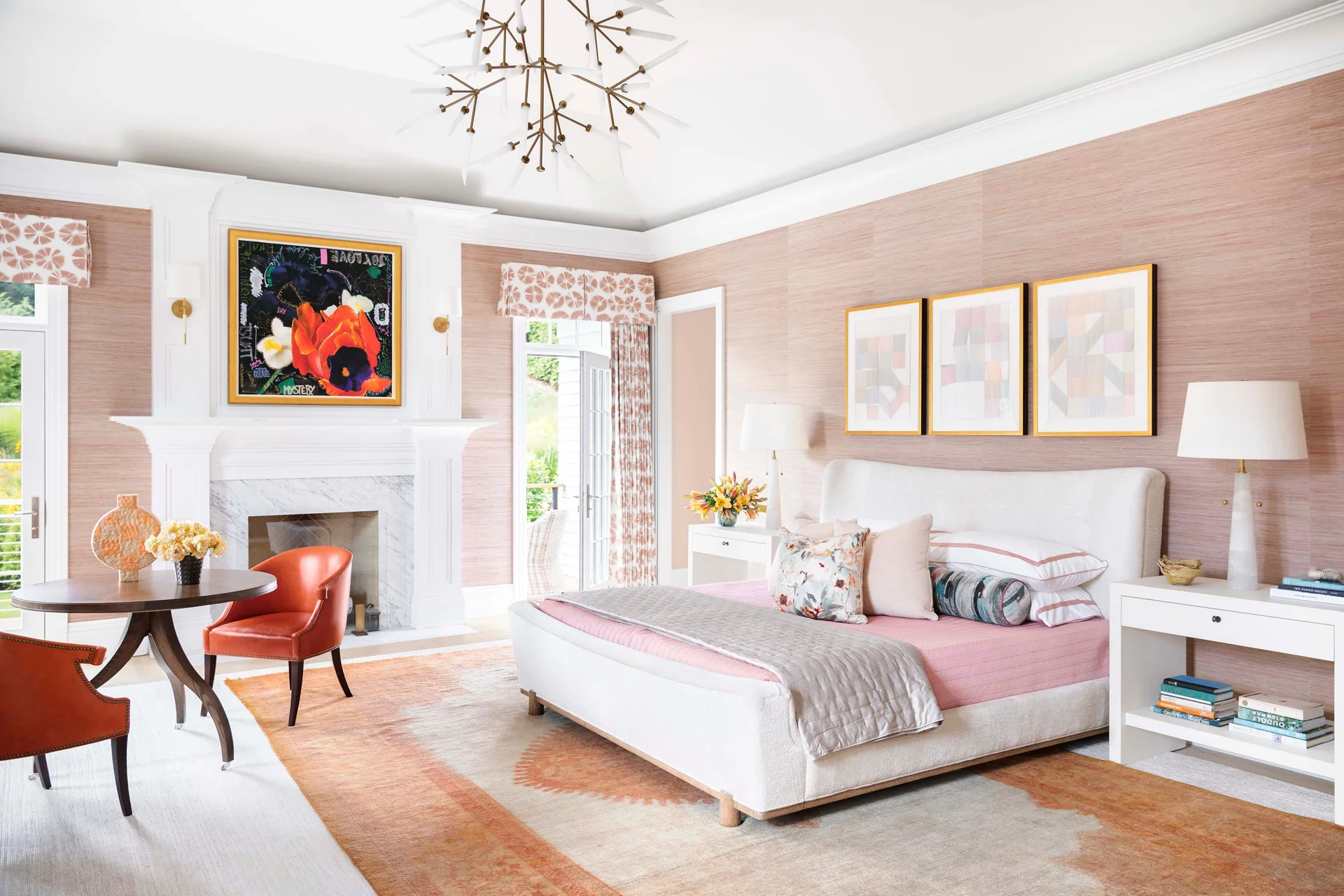 A bright transitional bedroom features pink grasscloth wallpaper, a white upholstered bed, a white marble fireplace, and orange leather armchairs.
