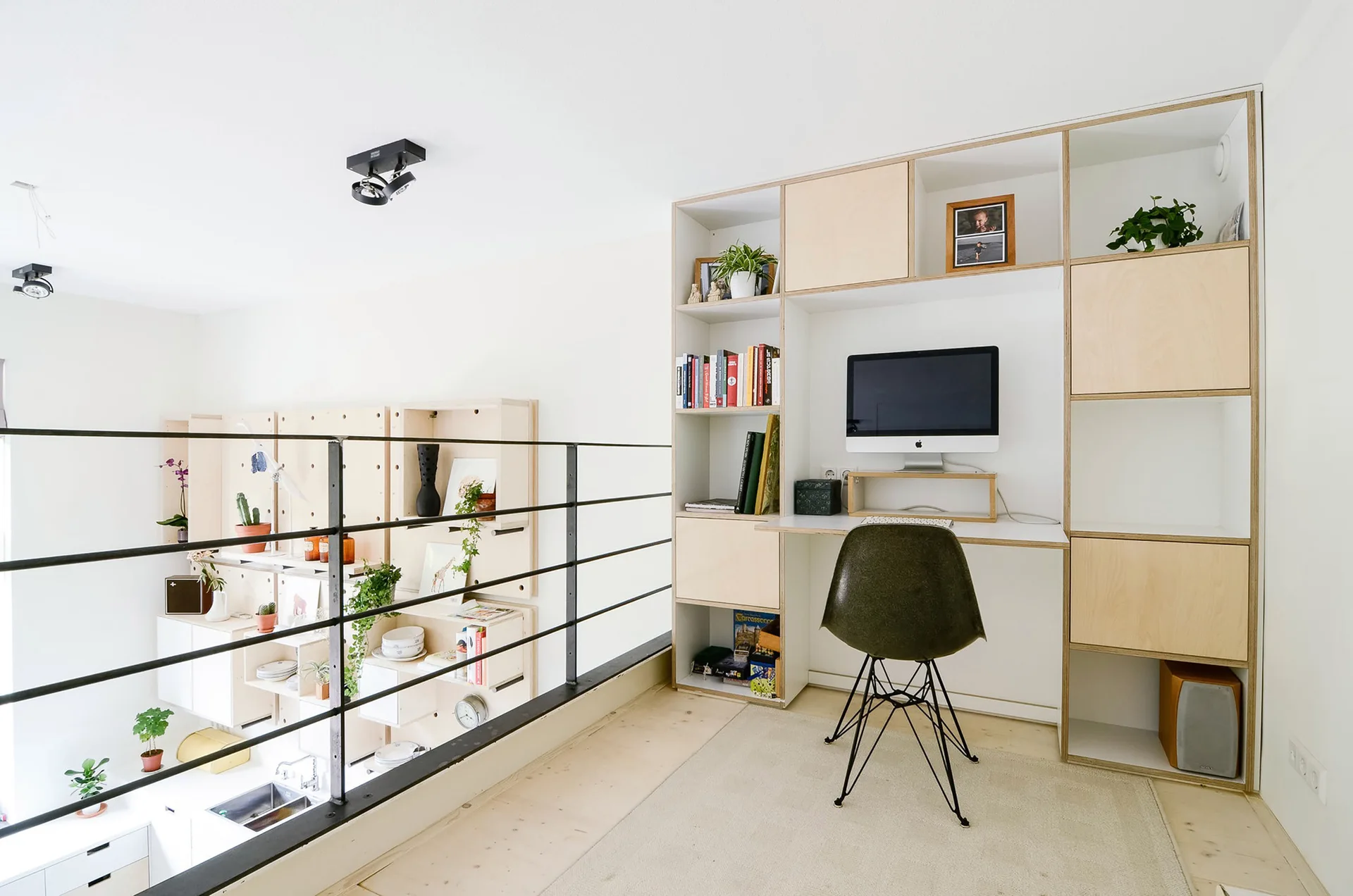 A minimalist home office on a mezzanine level with a built-in plywood desk and shelving unit next to a black safety railing.