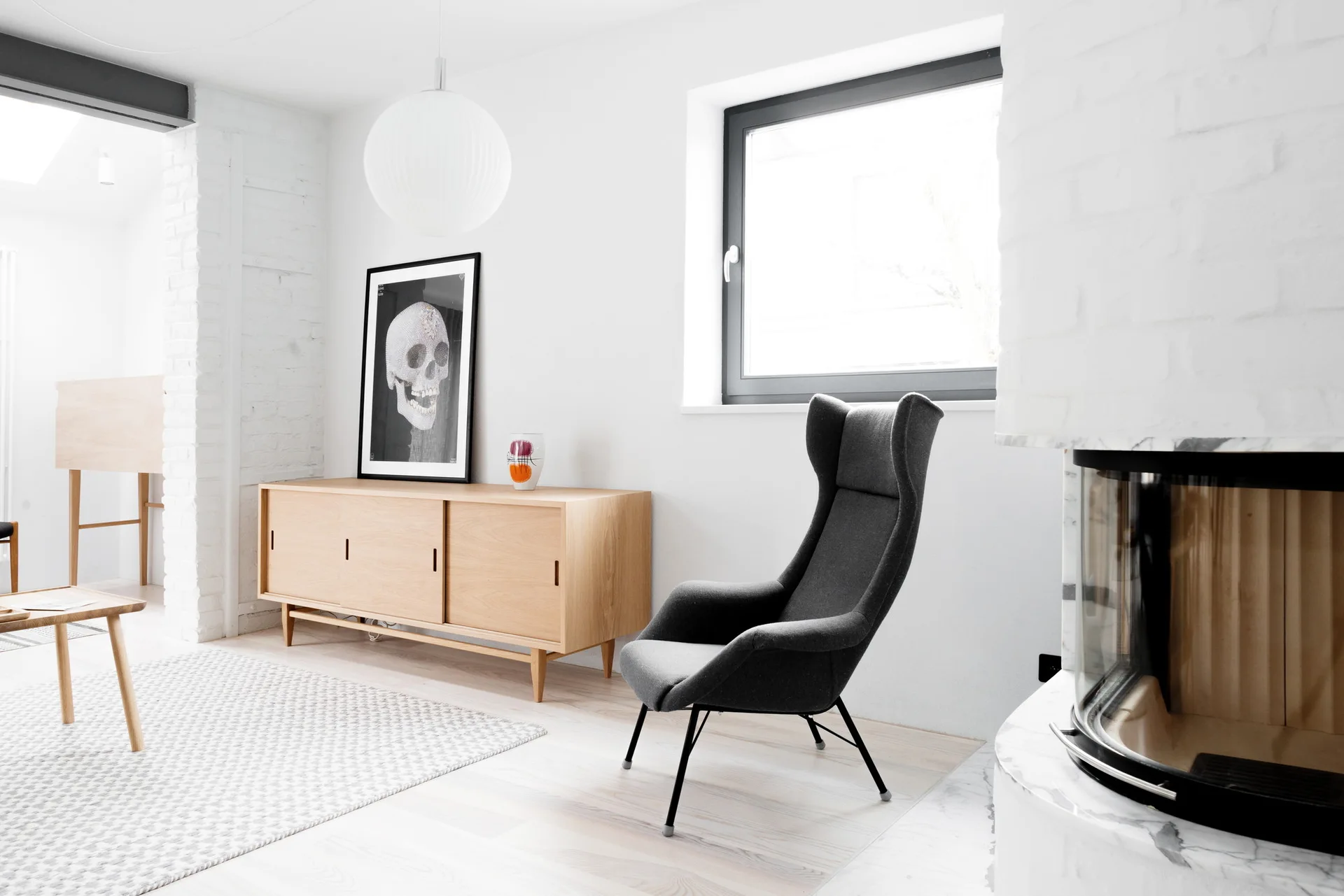 A minimalist living room features a charcoal high-back chair, a light wood sideboard with a framed skull print, and a modern fireplace.