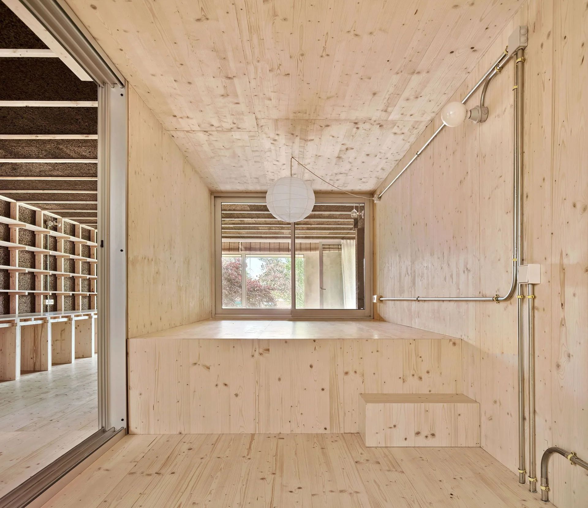 A minimalist room wrapped in light plywood featuring a raised platform, a paper globe pendant light, and exposed metal conduit on the wall.