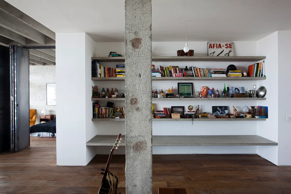 An industrial living room featuring thick built-in concrete shelves filled with books and decor, an exposed rough concrete column, and dark wood floors.