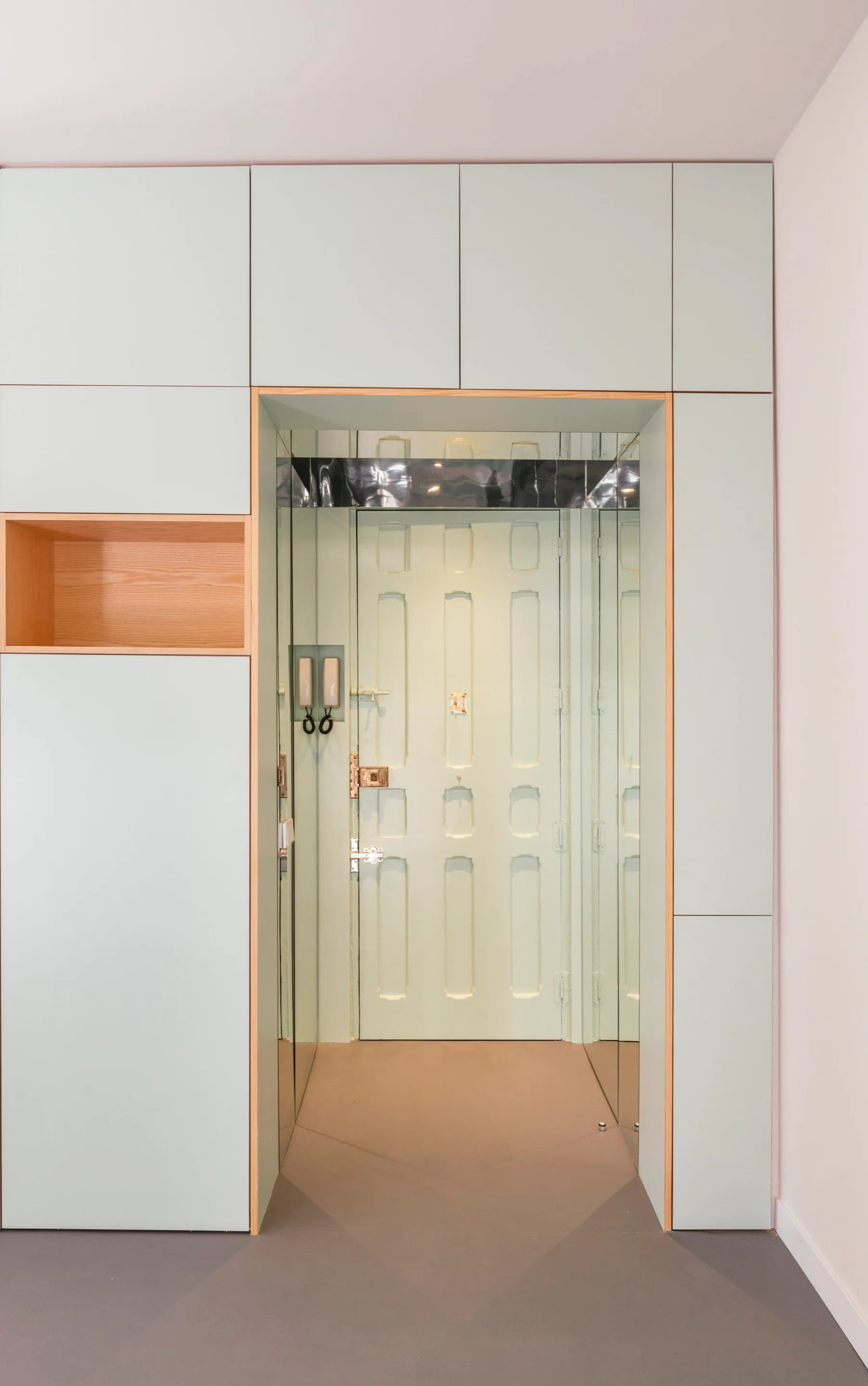 Matte pale green built-in cabinets surround a short entryway lined with mirrored walls that lead to a matching paneled door.