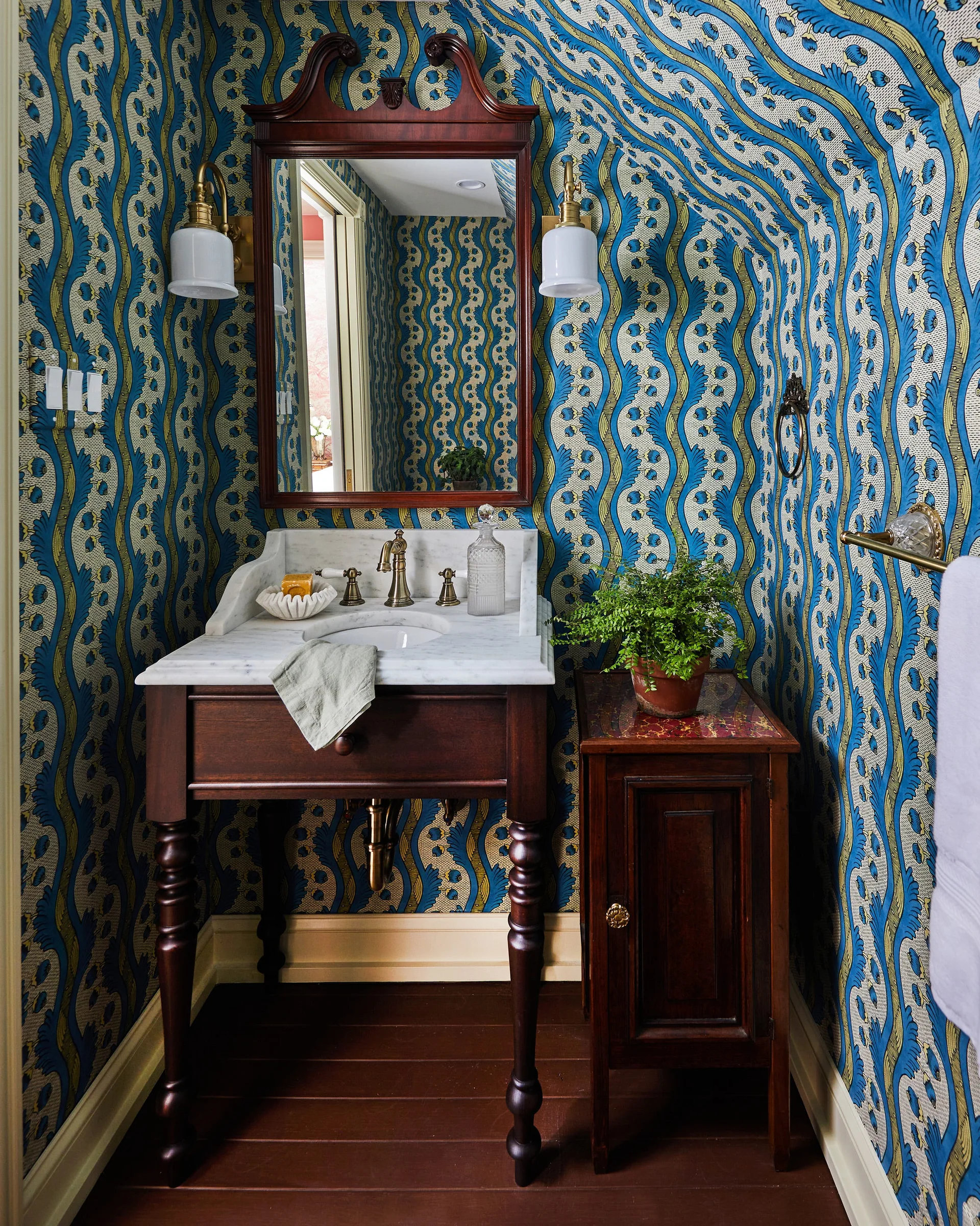 A powder room features bold blue and yellow patterned wallpaper, a dark wood vanity with a white marble top, and brass fixtures.