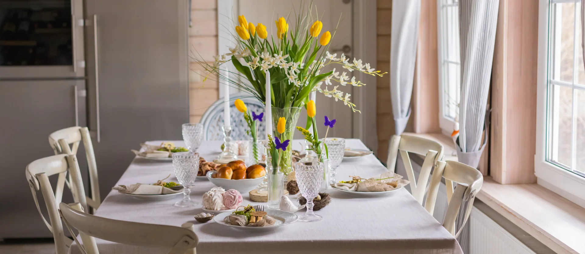 A set dining table next to a window features a white tablecloth, cut glassware, yellow tulips in a glass vase, and white wooden chairs.