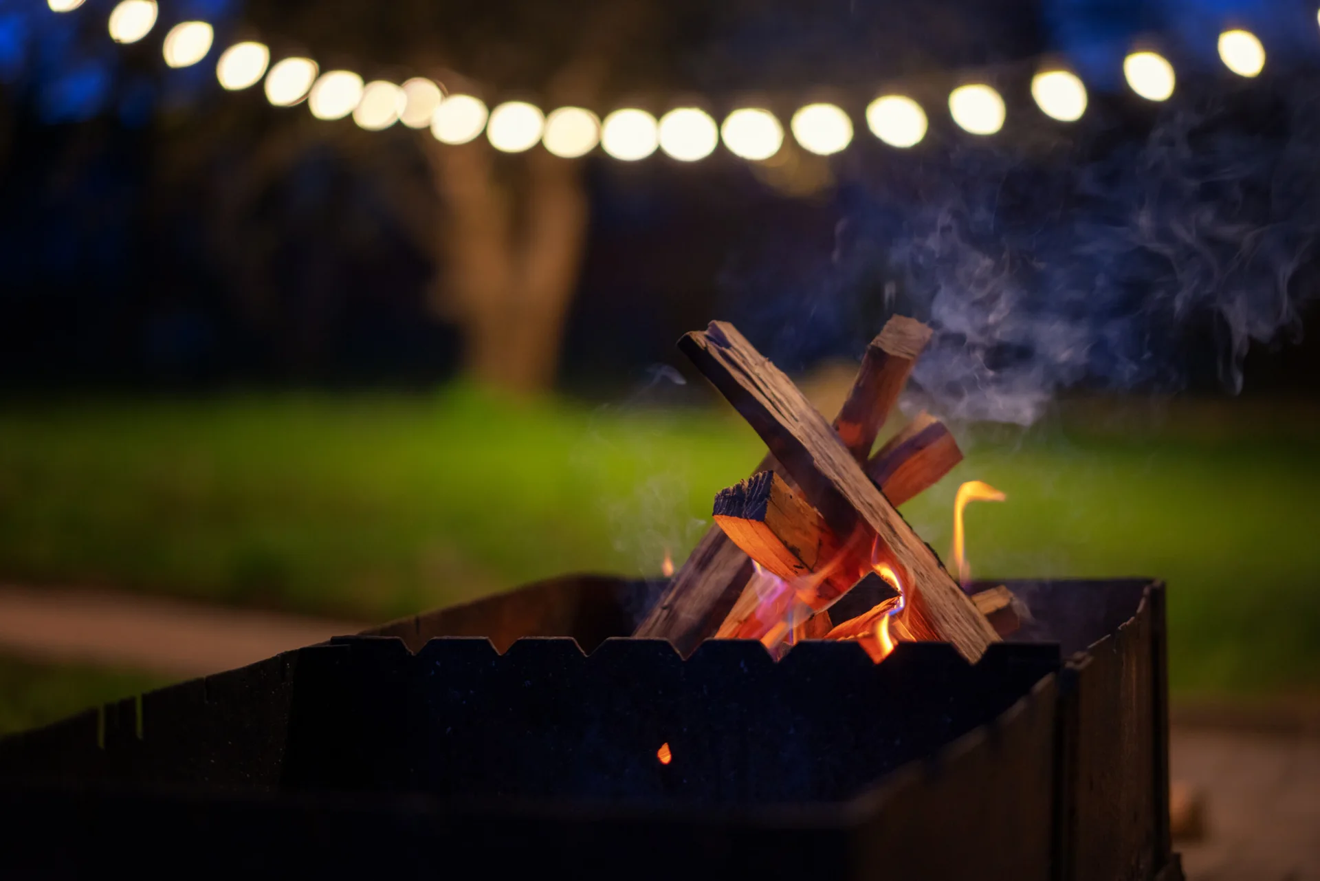 A black metal fire pit with burning wood logs stands on a lawn at night, backdropped by a curved string of glowing outdoor lights.