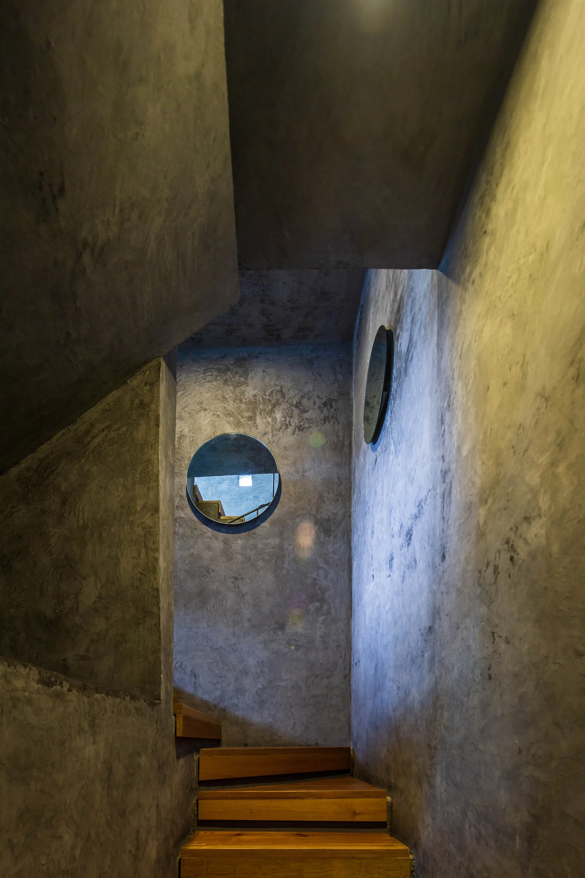 An enclosed minimalist staircase with warm wooden treads, textured grey concrete walls, and two round mirrors.