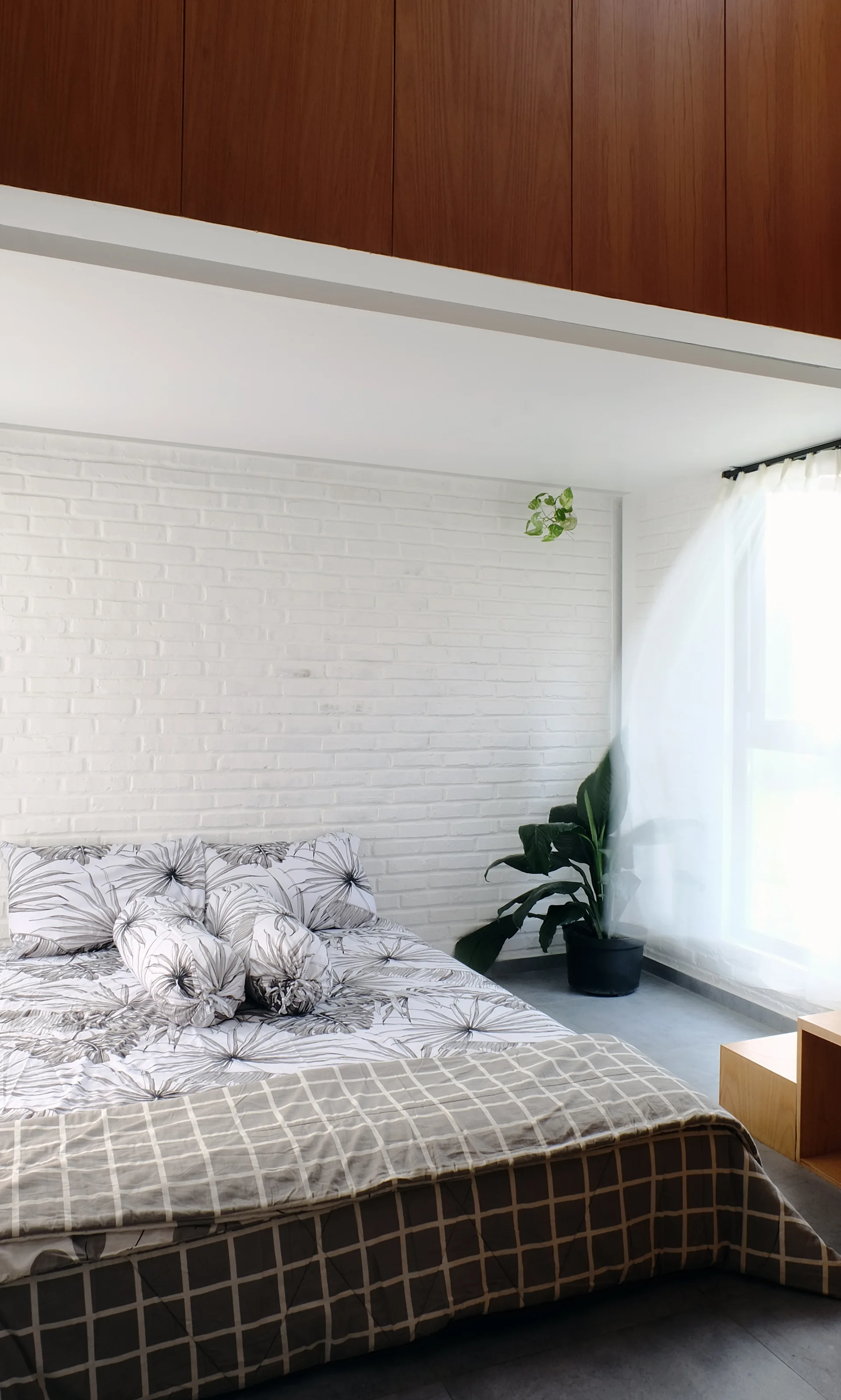 A modern bedroom features patterned botanical bedding on a bed against a white painted brick wall next to a sheer curtained window.