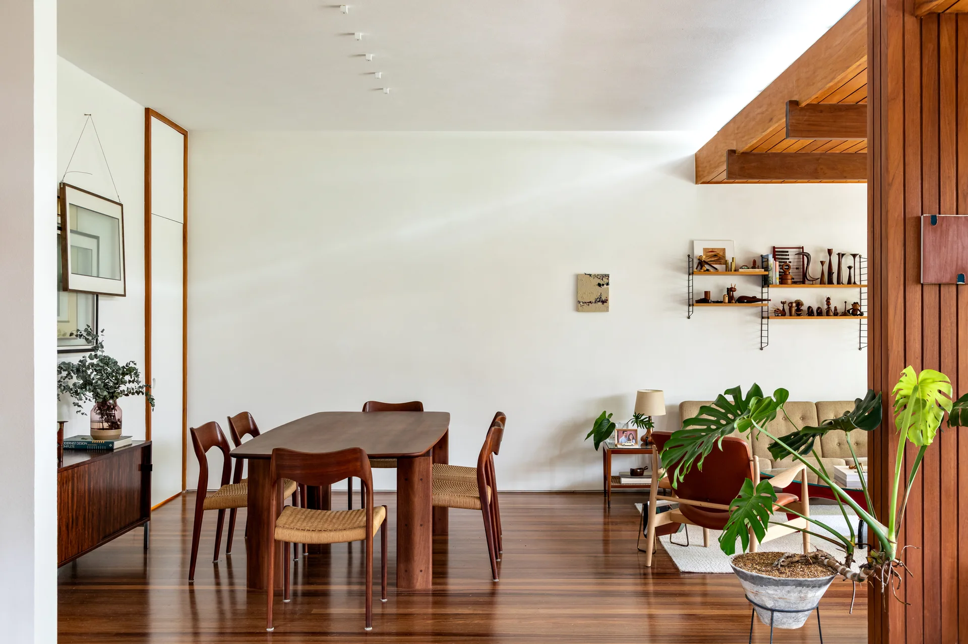 A mid-century modern dining room features a dark wood table with woven chairs, polished wood floors, and exposed ceiling beams.