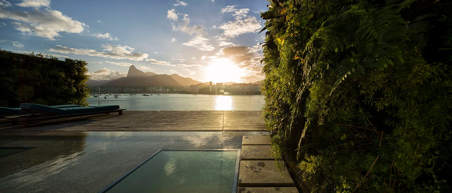 An outdoor pool terrace features wood plank decking, a lush green living wall, and sun loungers overlooking a bay at sunset.