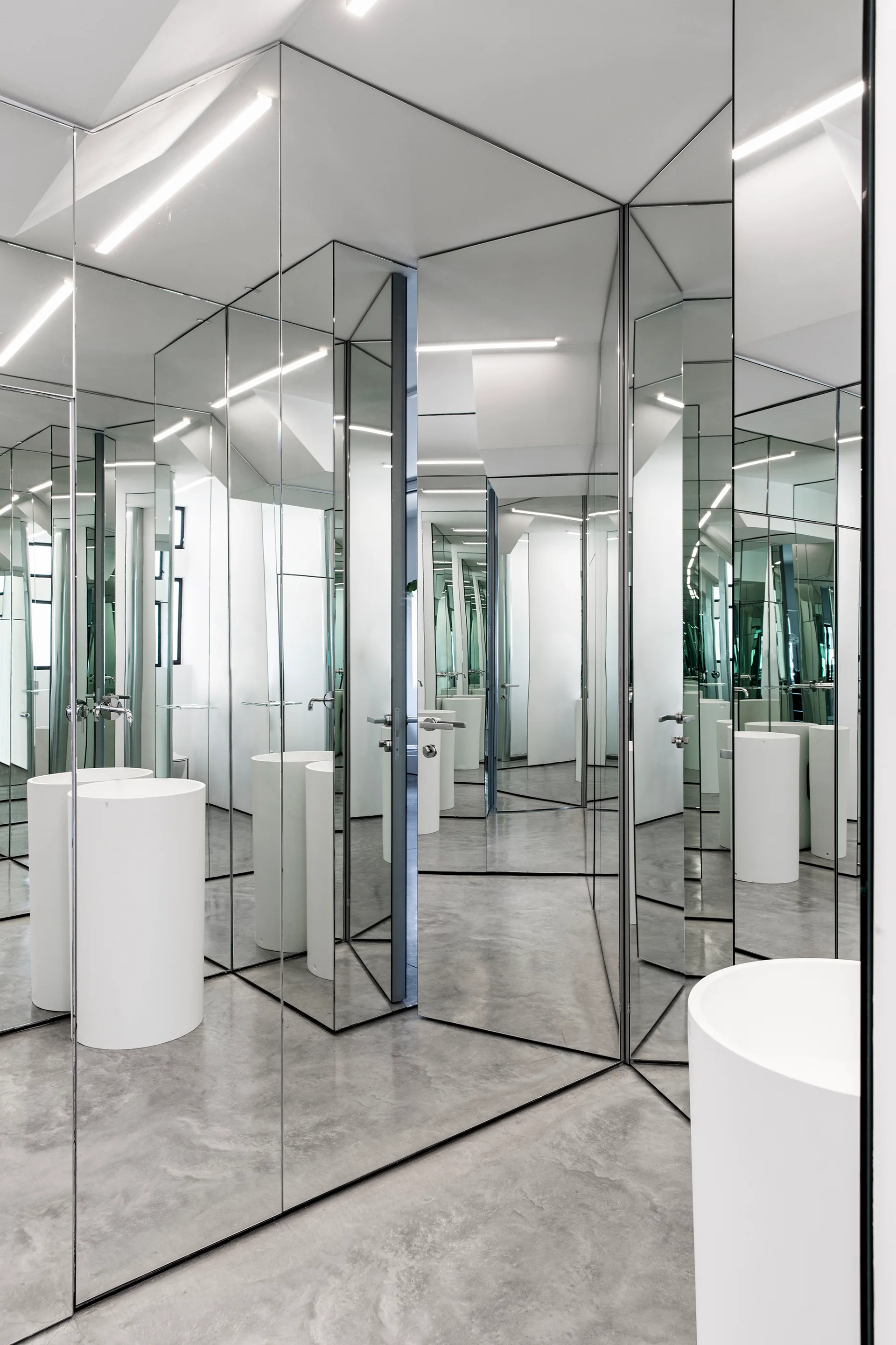 A minimalist bathroom features full-height mirrored walls reflecting white cylindrical pedestal sinks and a grey concrete floor.