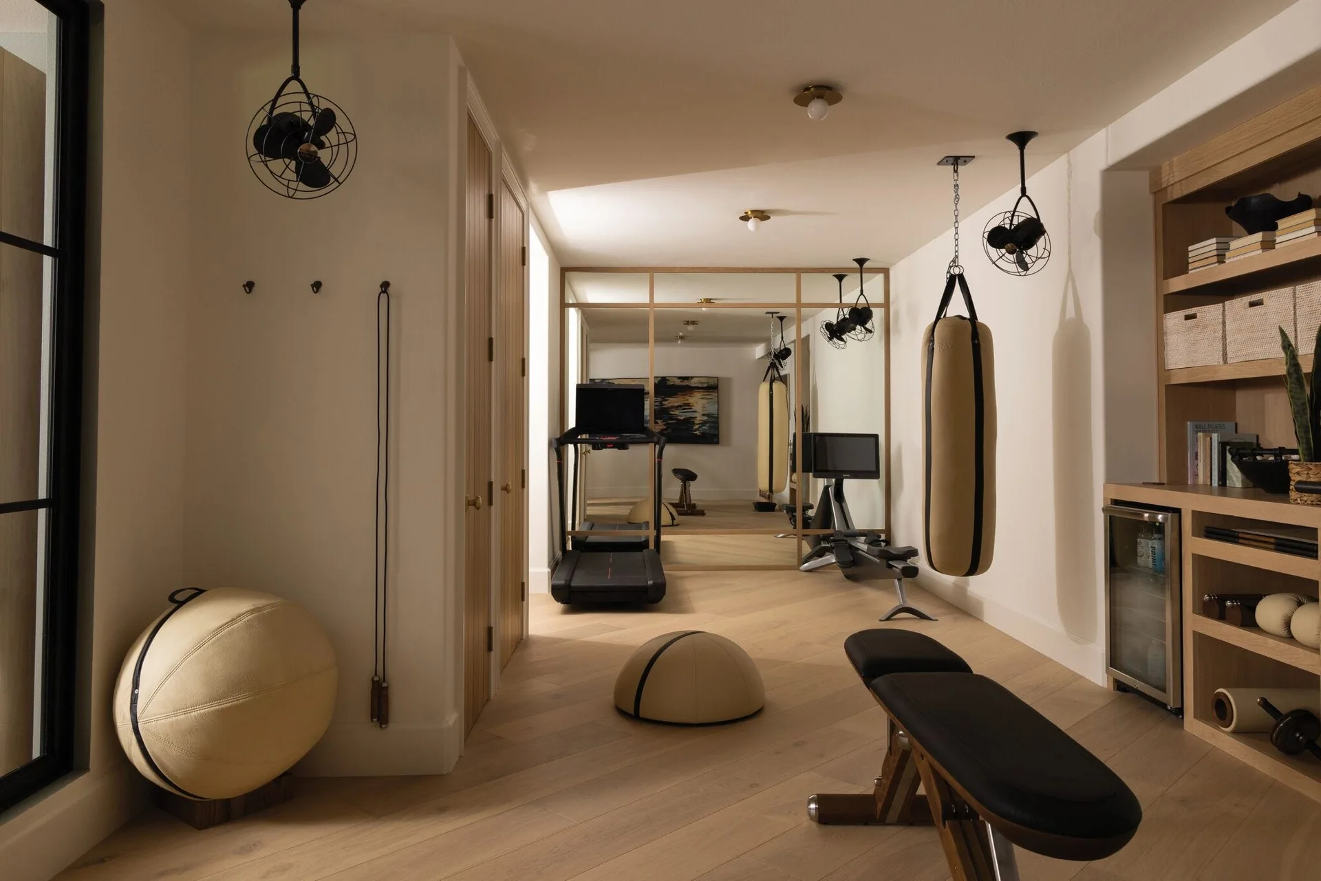 A modern home gym features light wood flooring, a treadmill, a tan hanging punching bag, and custom wooden shelving with stored fitness gear.