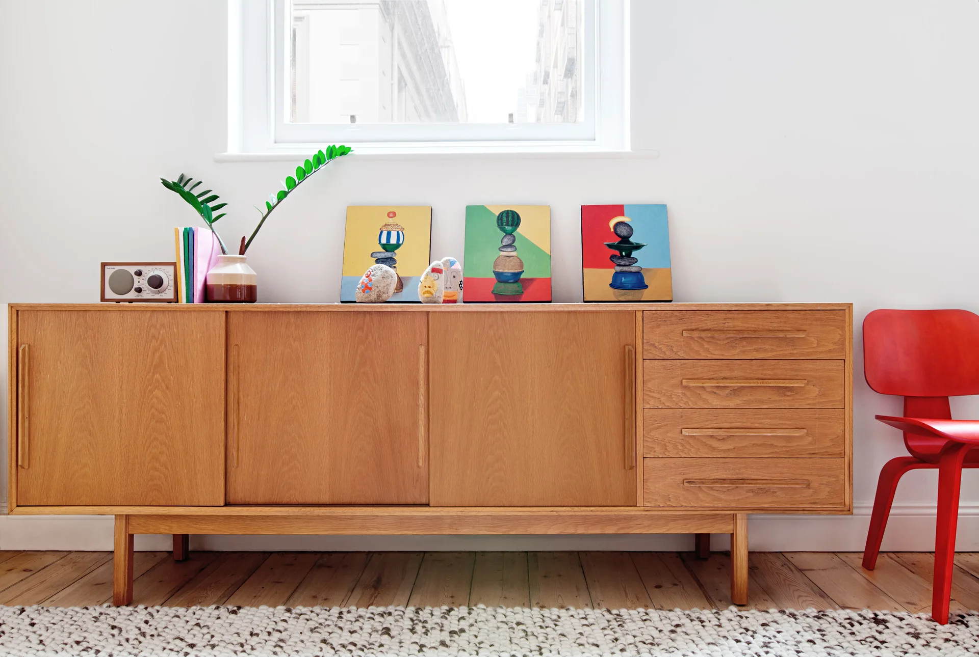 A natural wood mid-century modern sideboard with colorful art pieces sits on a textured rug next to a bright red wooden chair.