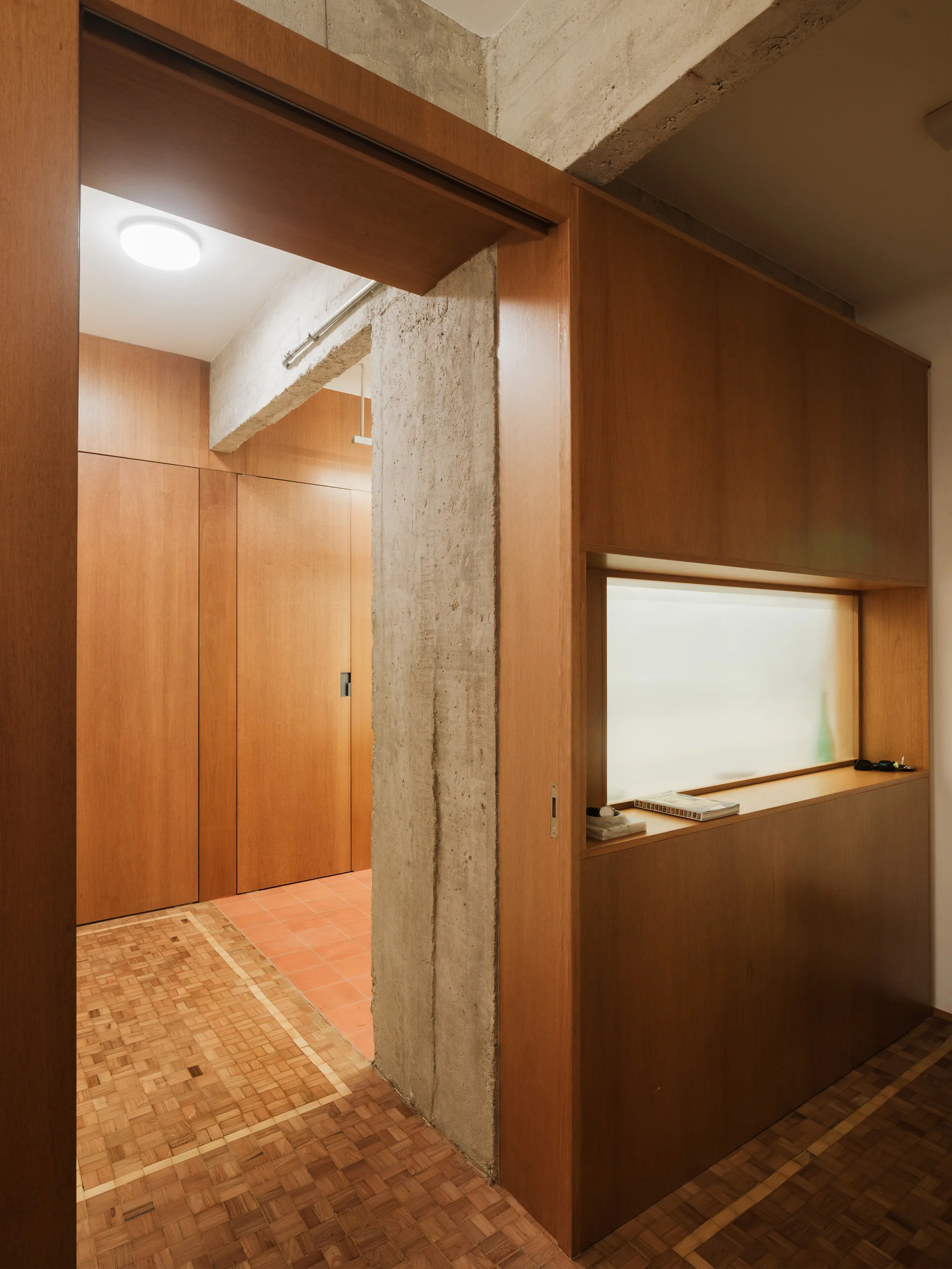 A minimalist hallway featuring warm wood wall panels, an exposed concrete pillar, a frosted glass window, and patterned parquet flooring.