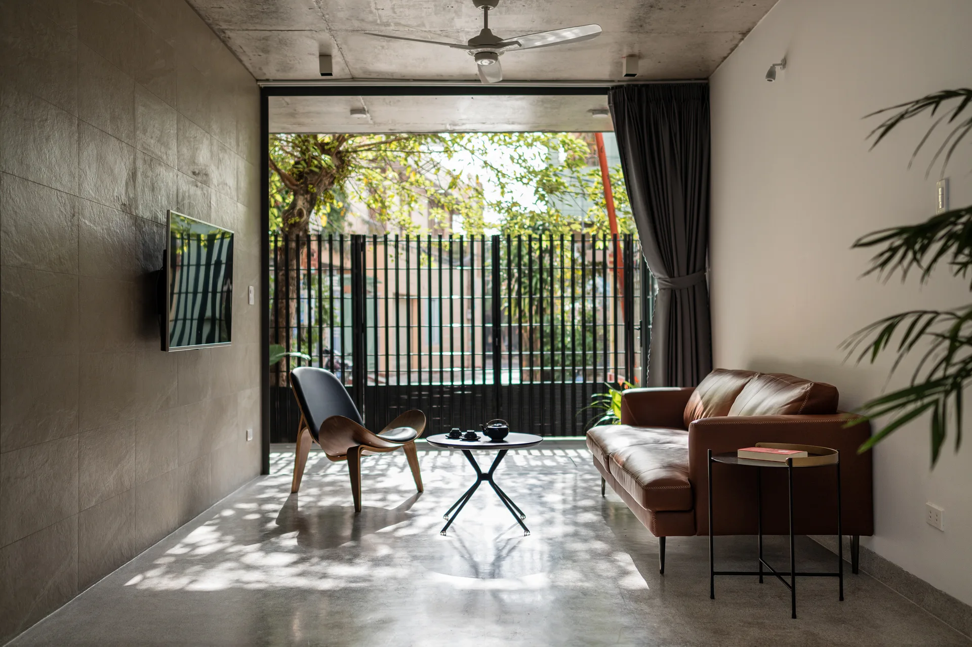 A modern living room with a brown leather sofa, wooden accent chair, and polished concrete floors opening onto an outdoor area with a slatted gate.