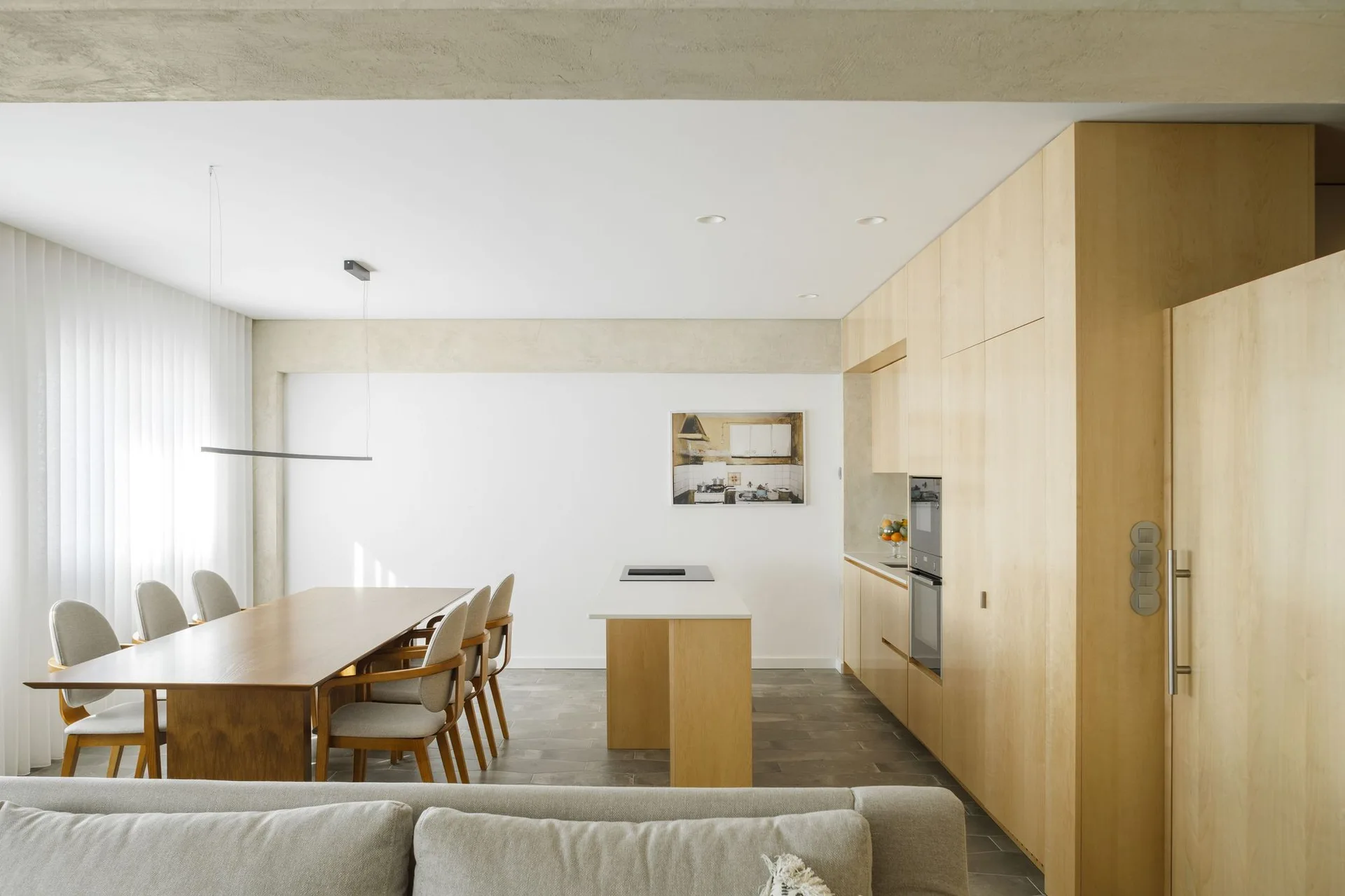 A minimalist kitchen and dining area features a light wood cabinet wall, a long wooden table, a simple island, and a grey tiled floor.