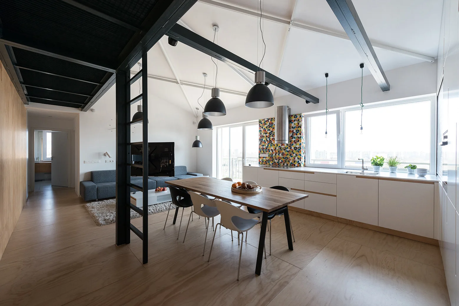 An open-concept loft kitchen features white cabinets, a geometric tile backsplash, and a wooden dining table under black steel ceiling beams.