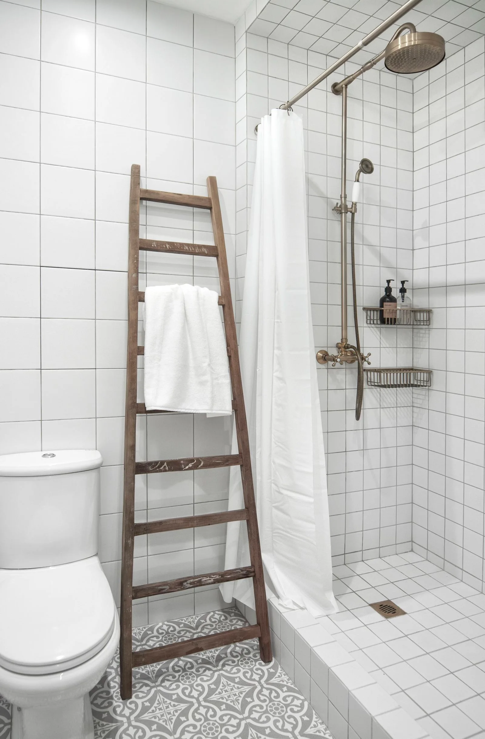 A bright bathroom features white square-tiled walls, grey patterned floor tiles, a white toilet, and a wooden towel ladder leaning against the wall.