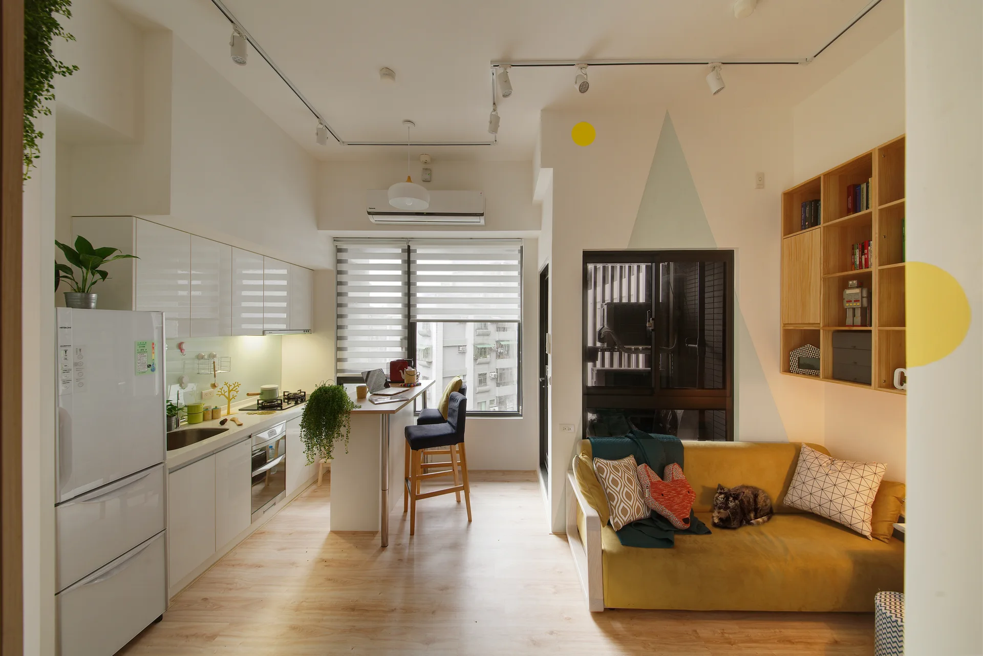 A compact open-plan room features a glossy white galley kitchen, a narrow breakfast bar with stools, and a small living area with a yellow sofa.