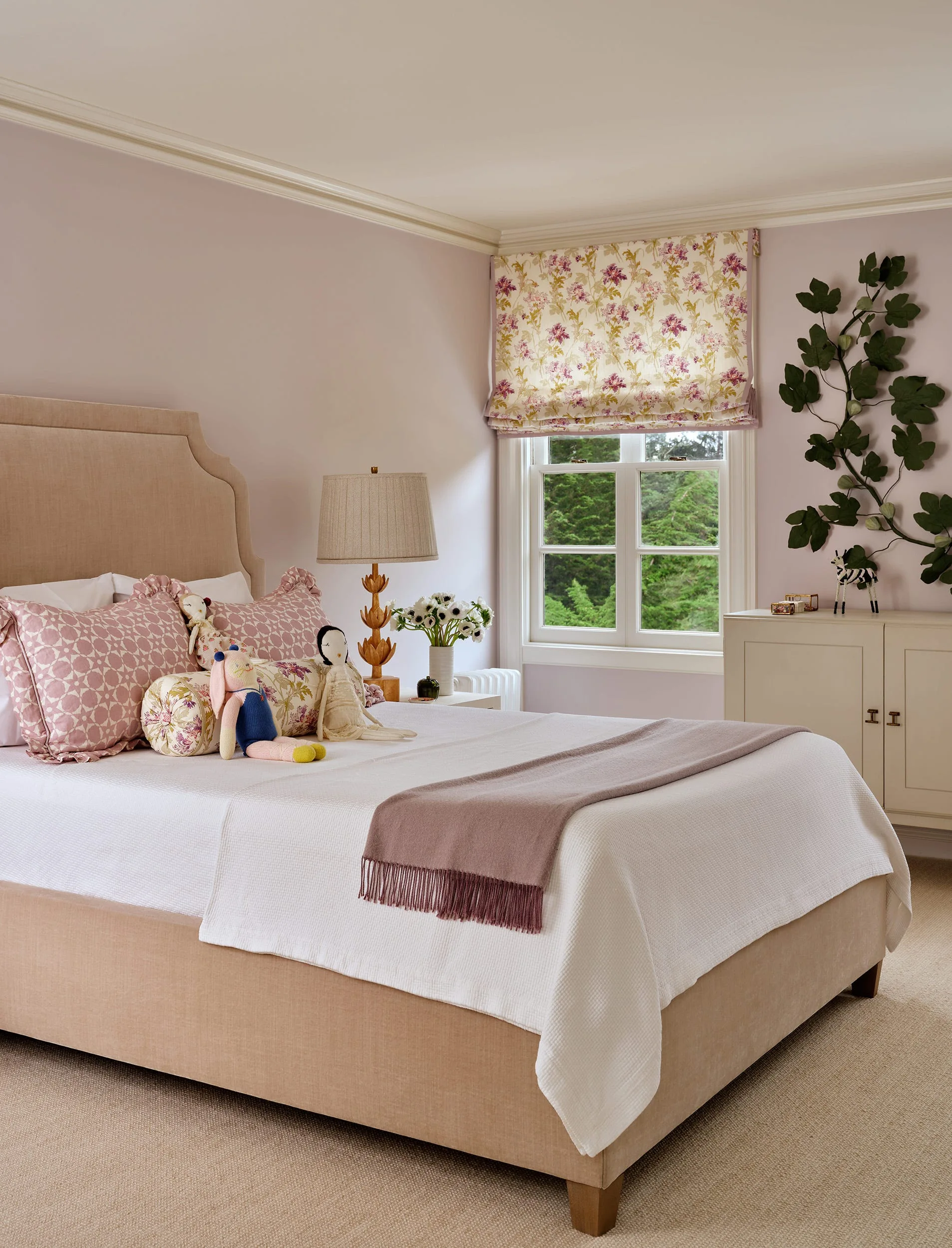 A bedroom with mauve walls, a tan upholstered bed with plush dolls, a floral roman shade over a window, and a green leafy wall sculpture.