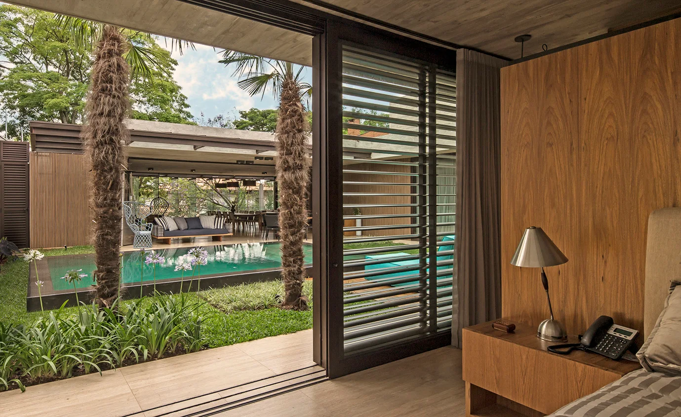 A modern bedroom with a wood-paneled partition wall opens through sliding glass doors to a landscaped pool area.