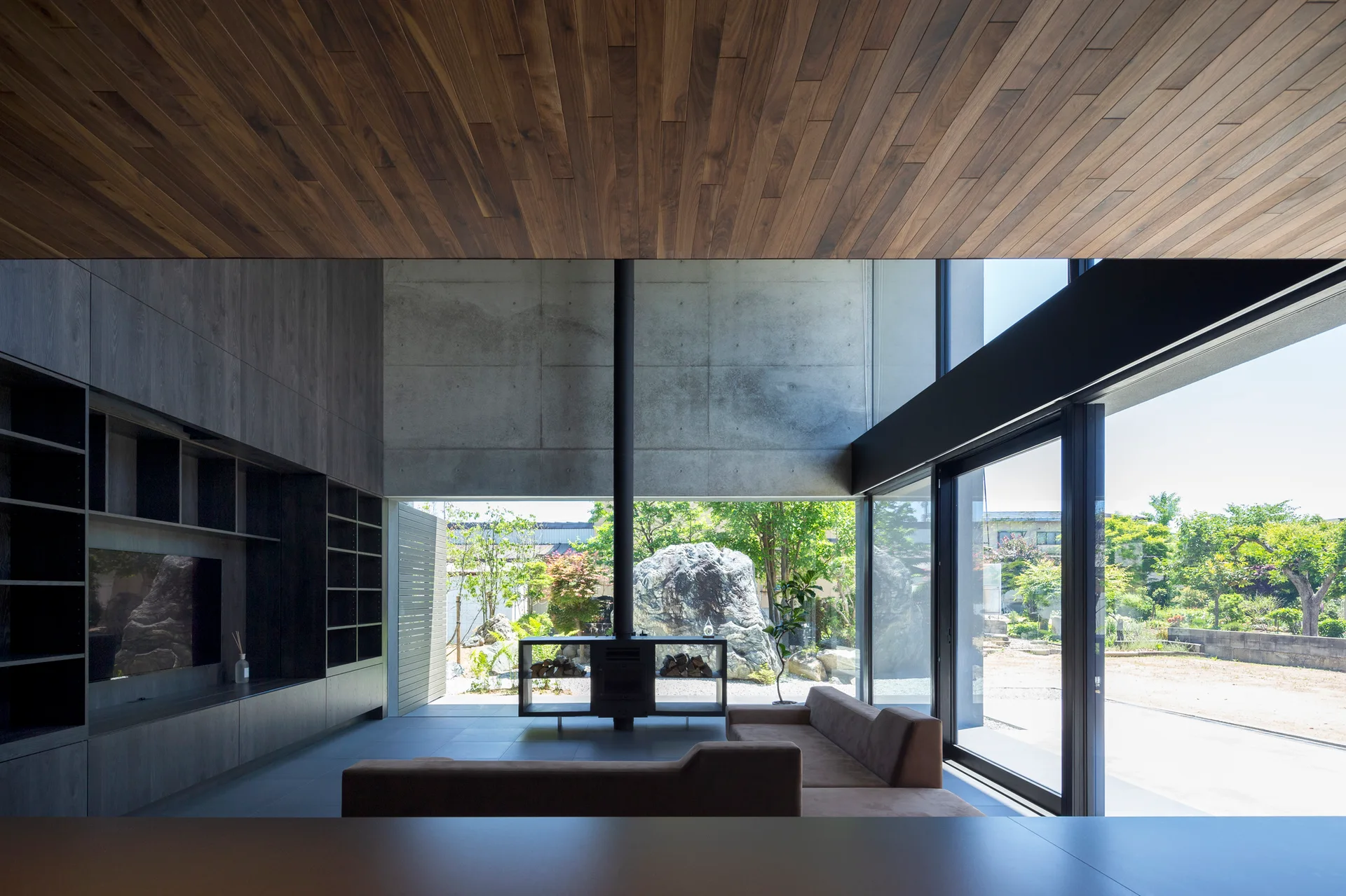 A minimalist living room features a wood plank ceiling, an exposed concrete wall, dark built-in cabinetry, and large sliding glass doors opening to a garden.
