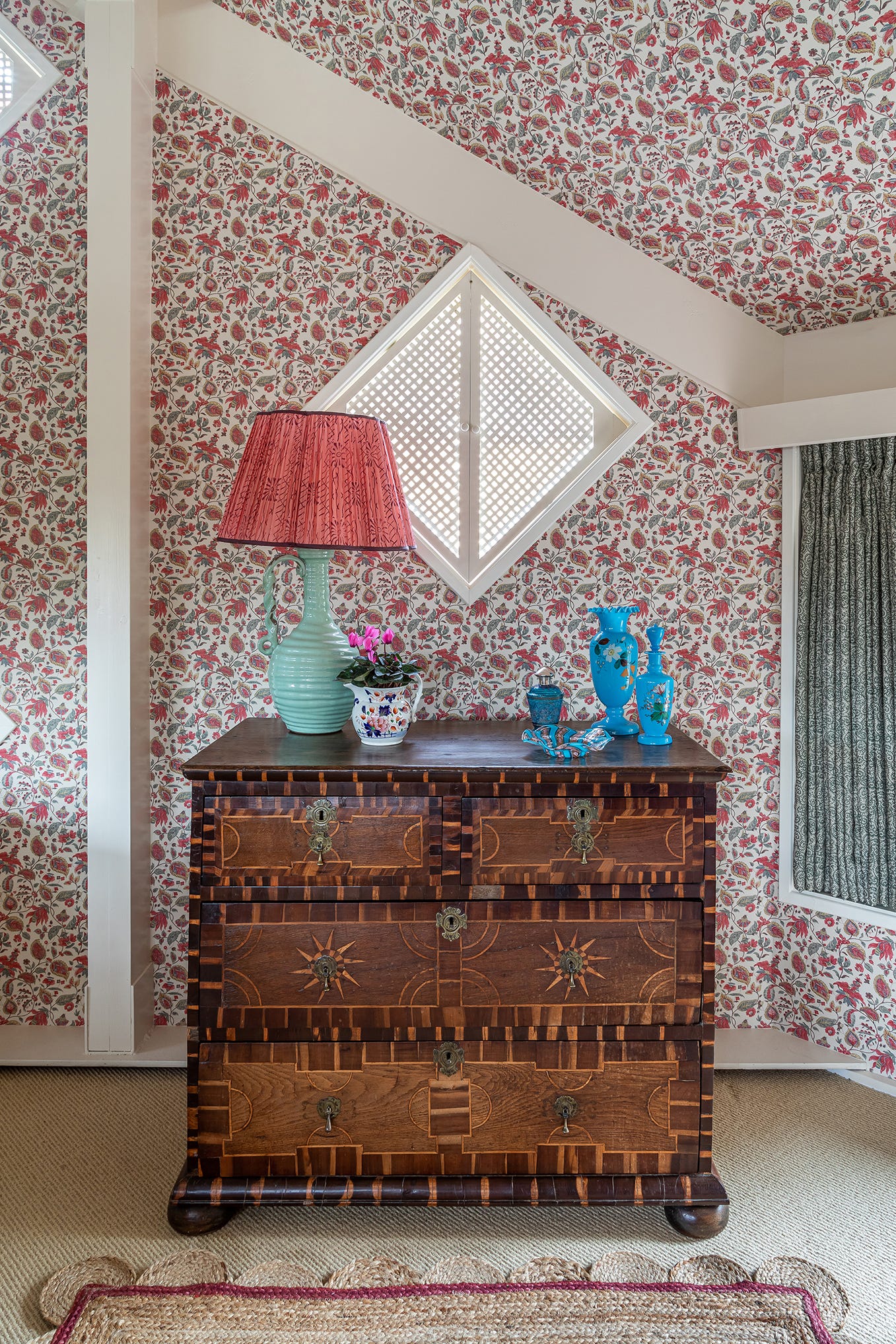 An antique inlaid wood chest of drawers stands against a wall covered in red floral wallpaper beneath a diamond-shaped lattice window.