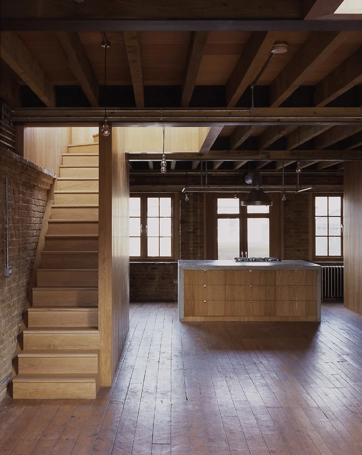 An industrial loft kitchen features a wooden staircase, an island with a concrete countertop, exposed ceiling joists, and a brick wall.