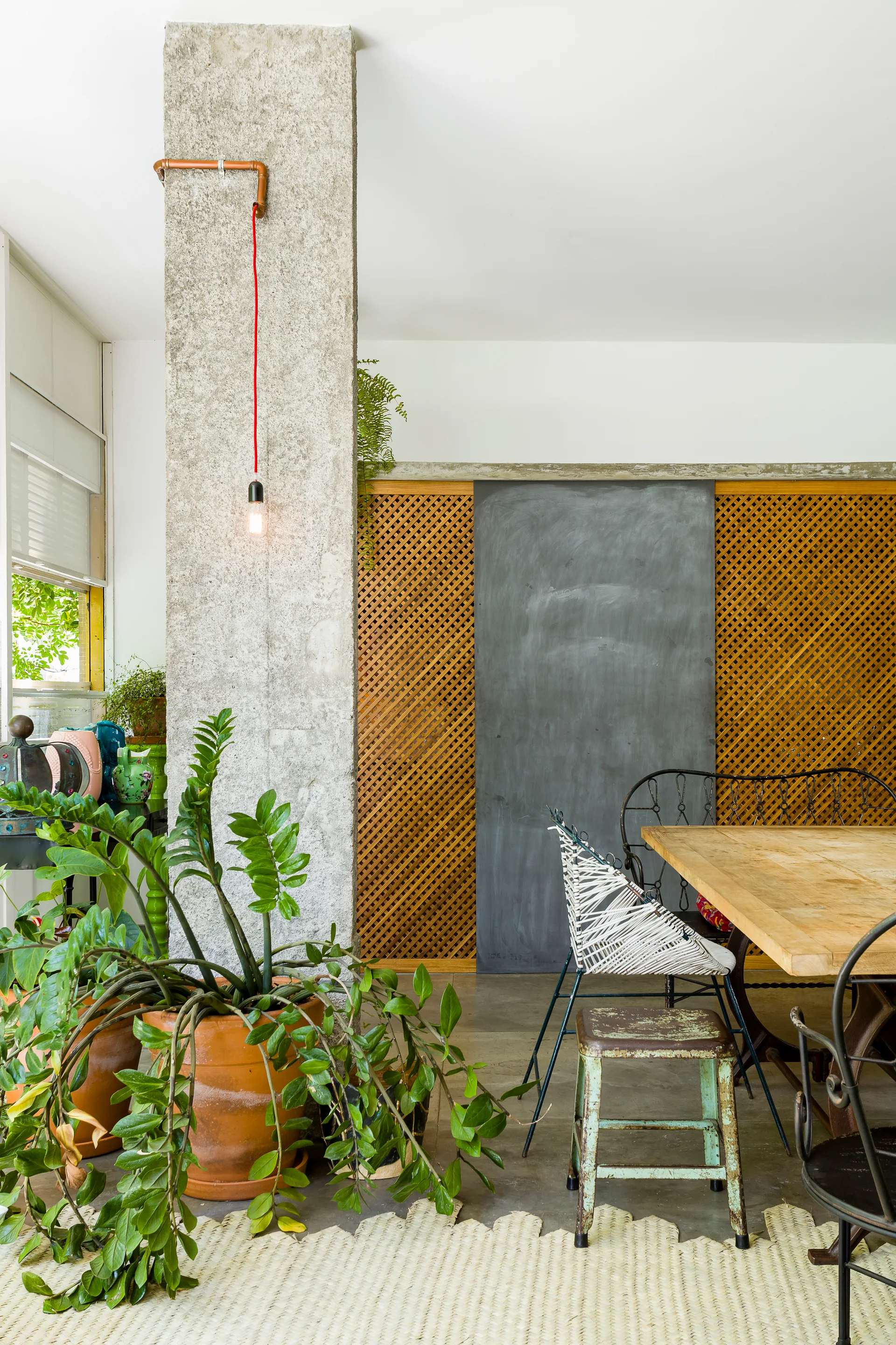 An interior dining room featuring an exposed concrete column with a red cord pendant light, a wooden table, and a decorative wood lattice wall panel.