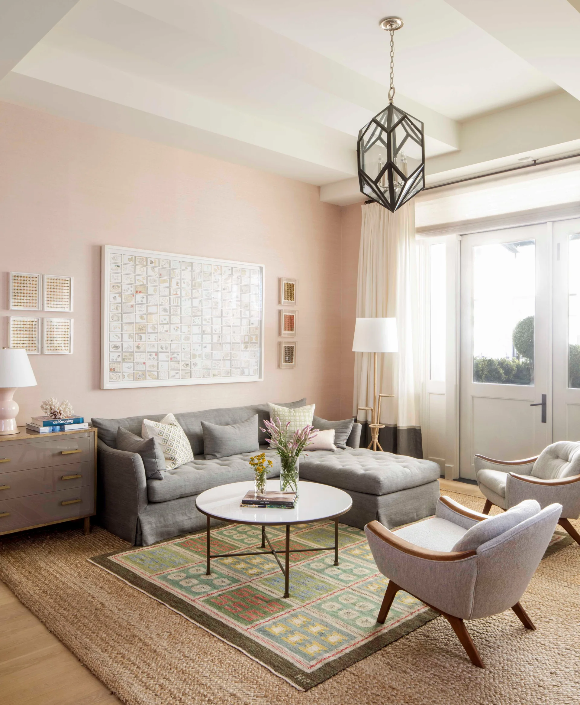 A living room with blush walls features a grey sectional sofa, two mid-century armchairs, a patterned rug layered on sisal, and a black geometric pendant.