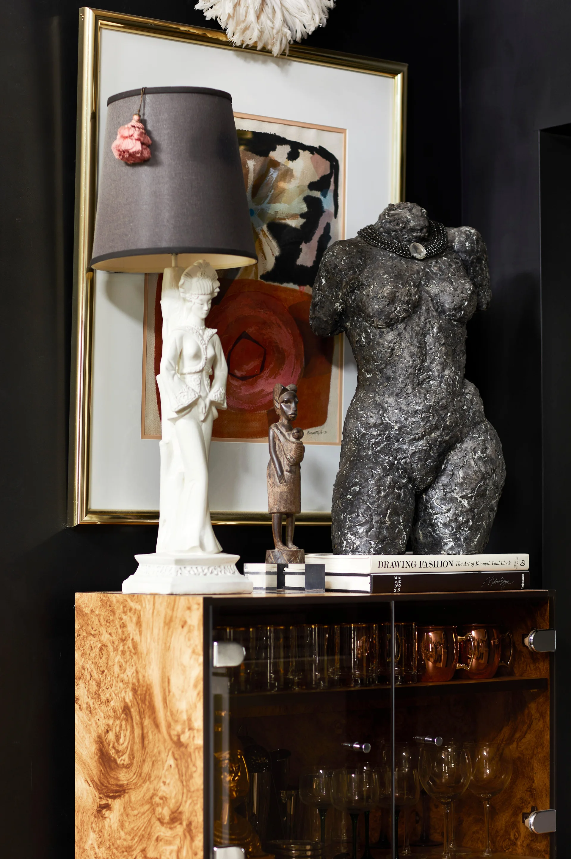 An eclectic display atop a burl wood cabinet featuring a geisha-shaped lamp, a textured stone torso sculpture, and framed art against a black wall.
