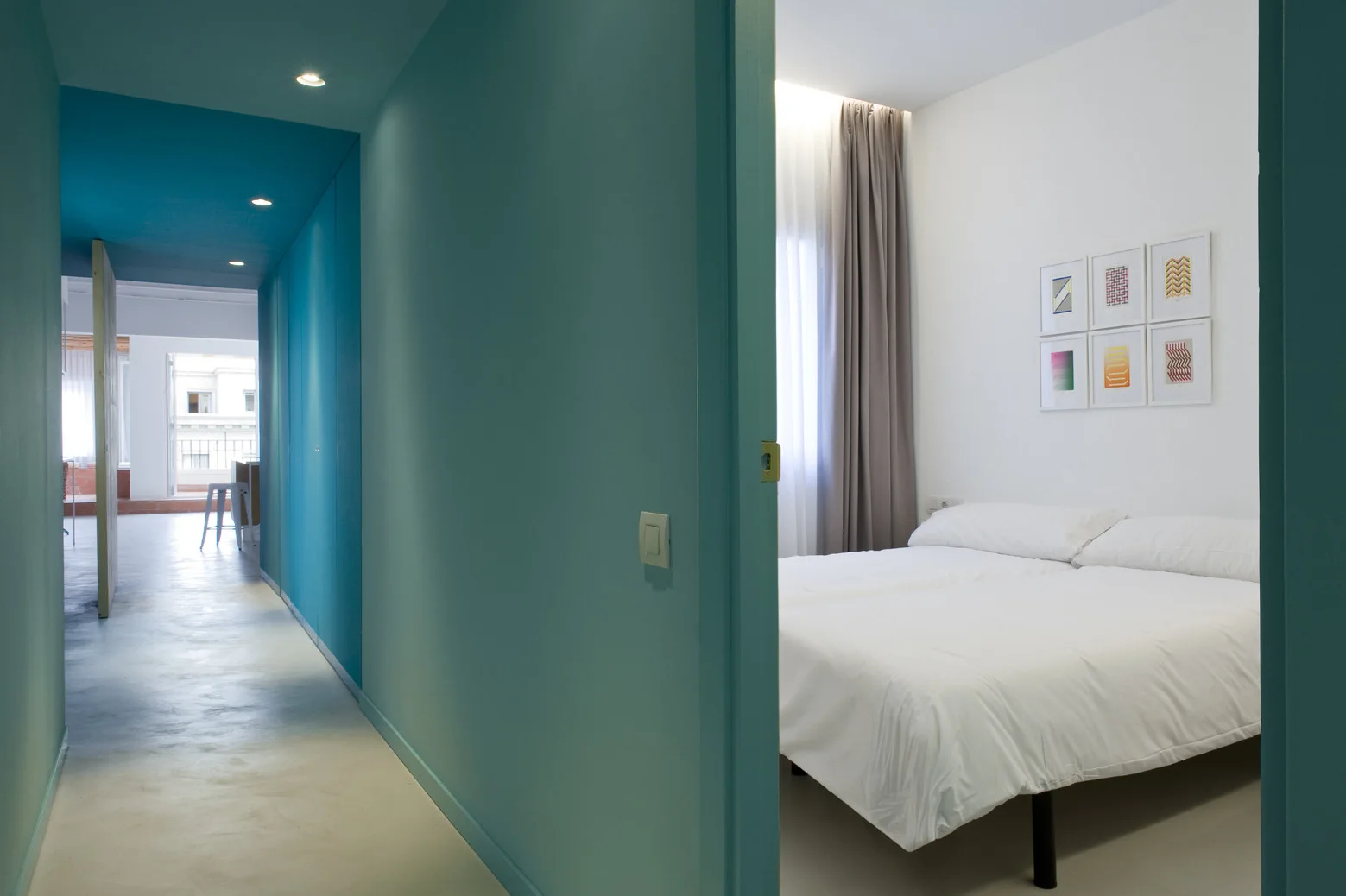 A view from a blue painted hallway with recessed lighting looking into a white minimalist bedroom with a bed and framed wall art.
