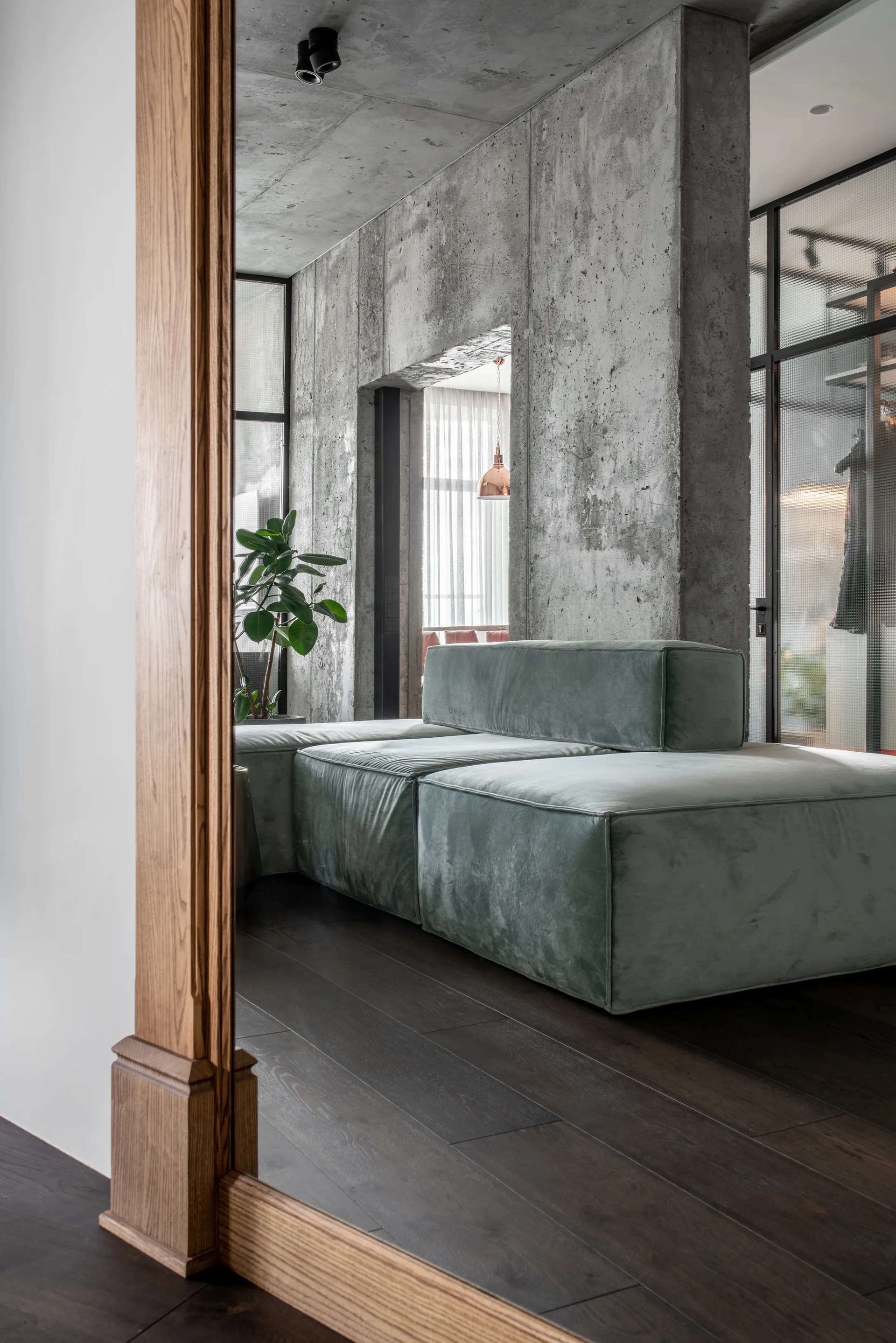 A modular sage green velvet sectional sofa sits against a raw concrete wall on a dark wood floor.