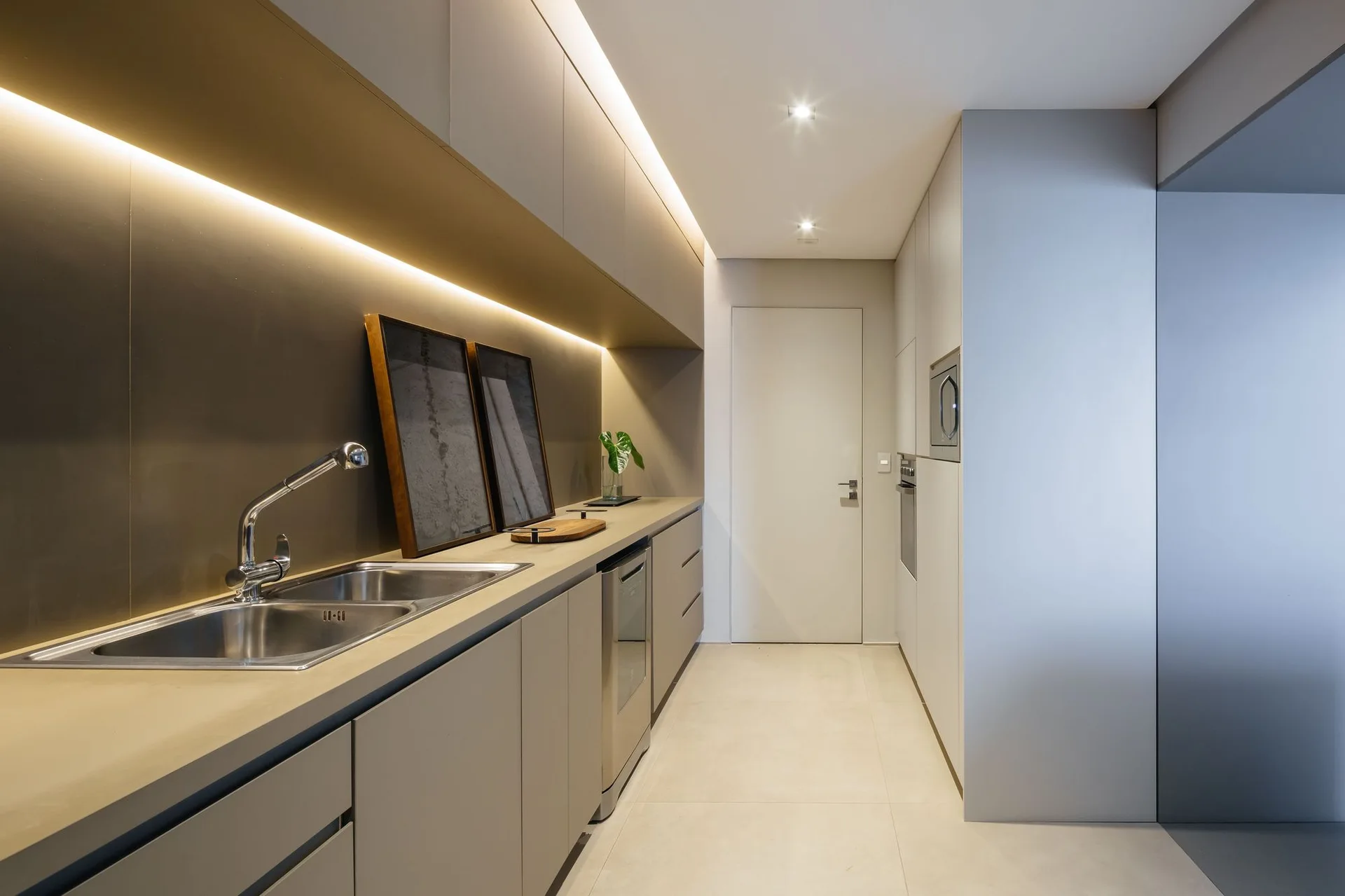 A modern galley kitchen features matte grey handleless cabinets, a stainless steel sink, built-in appliances, and warm LED strip lighting under the upper units.