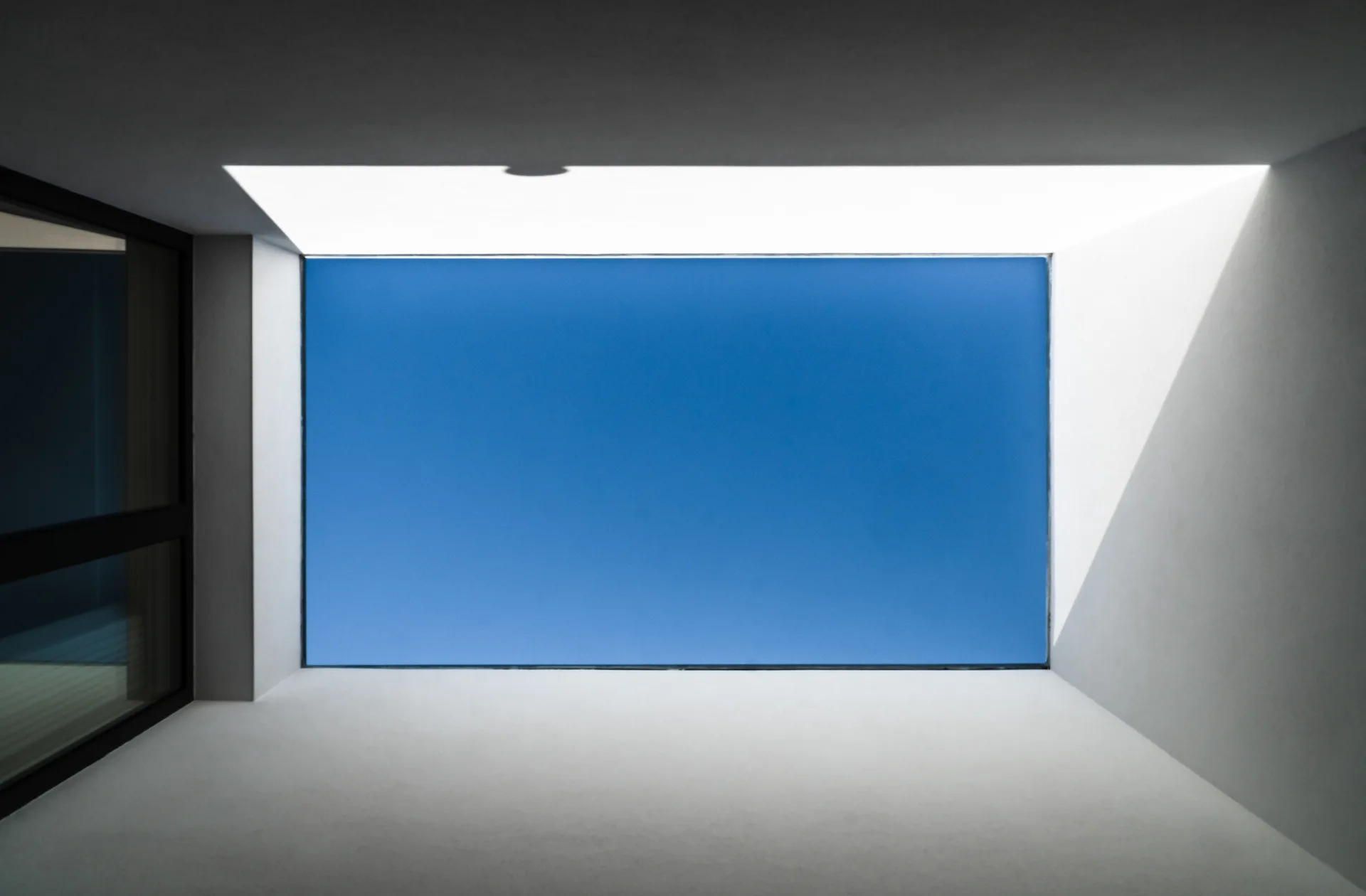 A minimalist white ceiling features a large rectangular skylight looking up at a clear blue sky, flanked by smooth plaster-like walls.