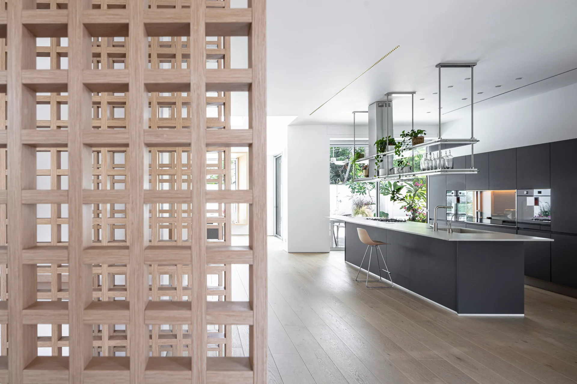 A modern kitchen features a charcoal grey island and cabinetry, a hanging metal shelf, and a light wood grid partition in the foreground.