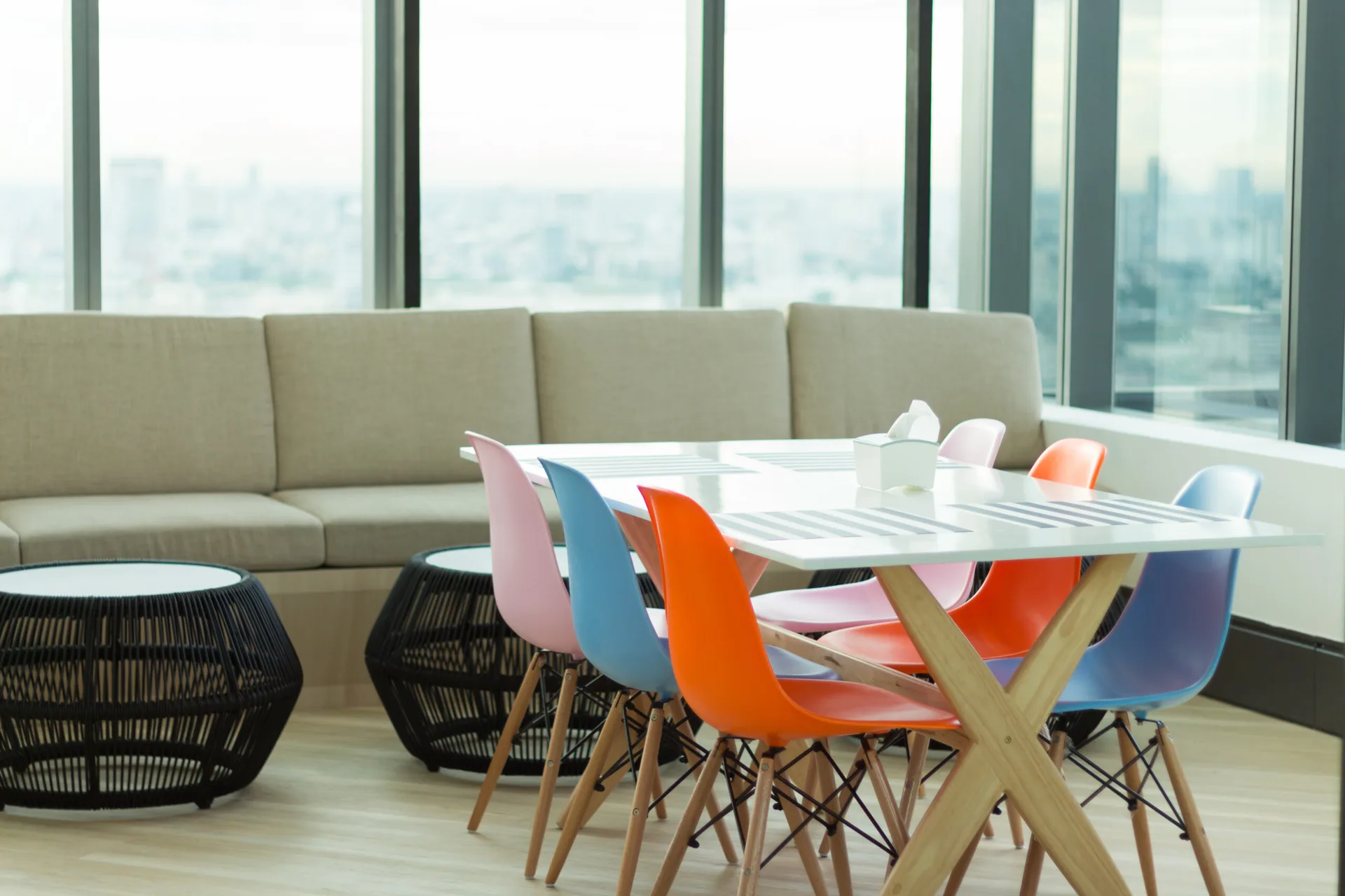 A modern white dining table surrounded by colorful plastic chairs in pink, orange, and blue near large windows.