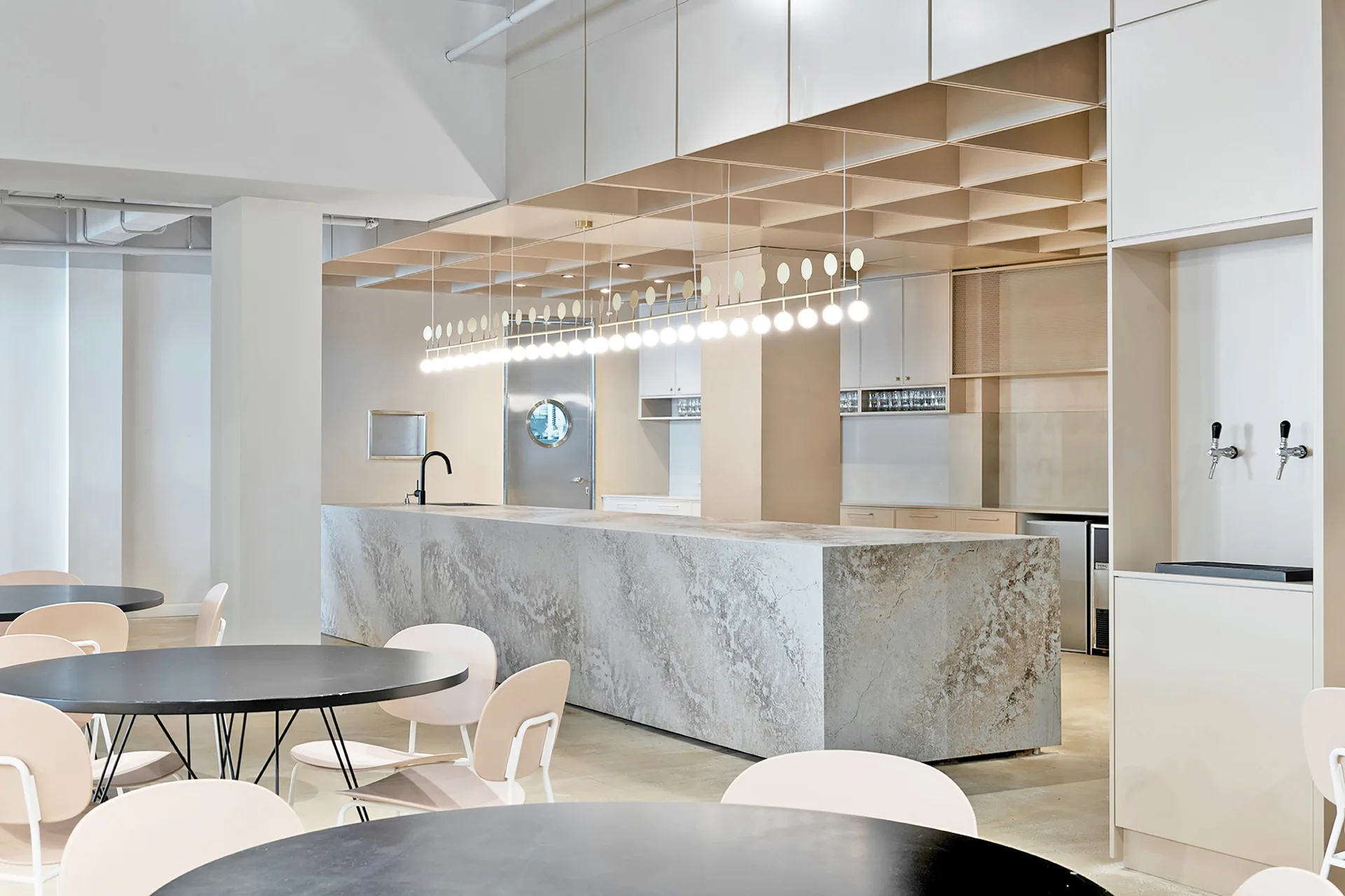 A modern cafe interior features a long stone-like bar counter under a wooden ceiling grid and a linear pendant light, with circular tables in the foreground.